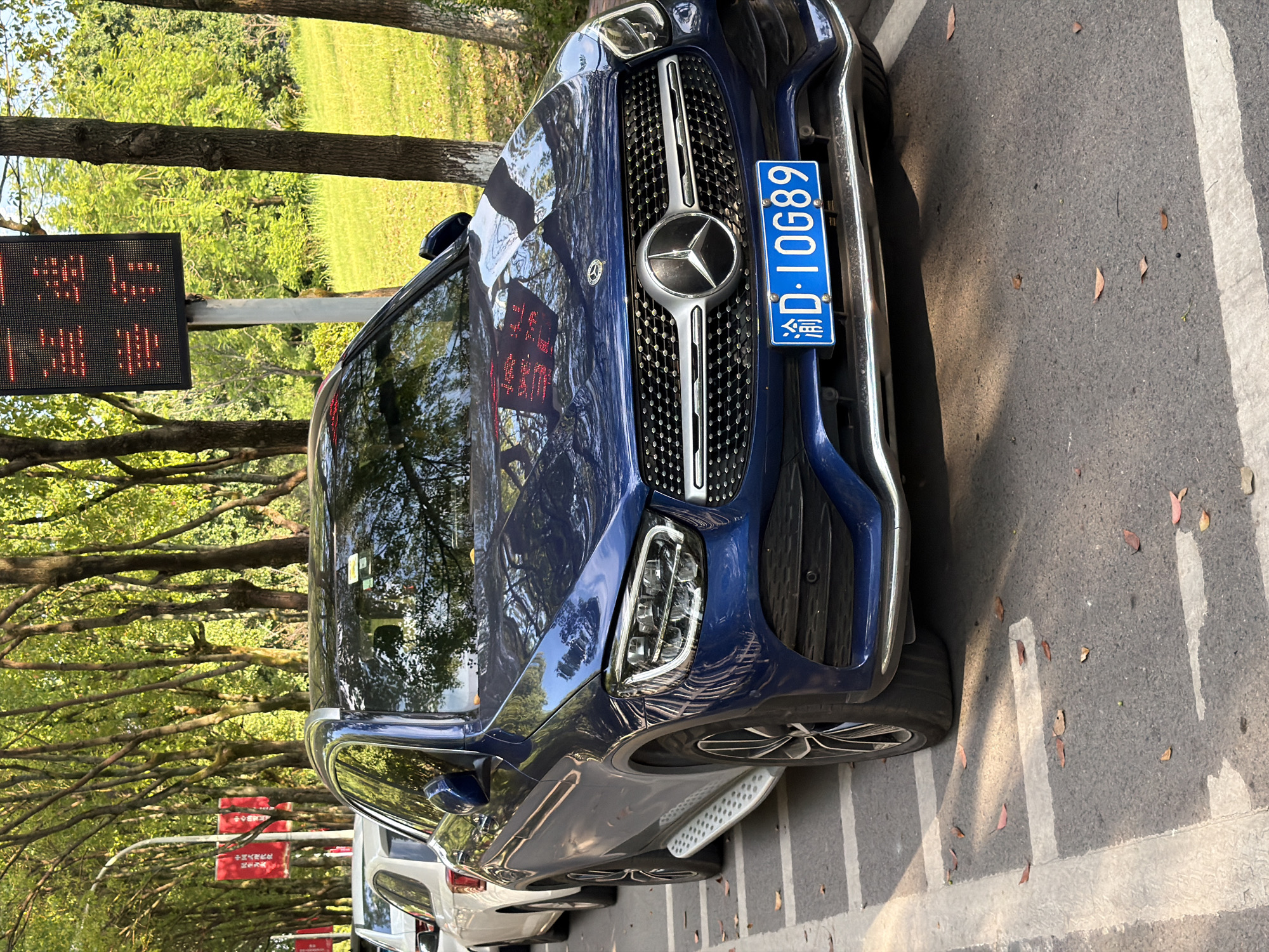 A dark blue Mercedes-Benz SUV is parked in a marked parking space on a paved road. The vehicle has a license plate that reads "渝 D·1OG89". To the left of the SUV, a white car is also parked. Behind the vehicles, a row of trees with green foliage lines the road, and a grassy slope is visible beyond the trees. A pole with a digital display showing red characters stands next to the SUV. Red banners with Chinese characters are hanging from poles along the side of the road in the background. The scene appears to be in an urban environment in Jiangjin, China, given the license plate prefix "渝" which corresponds to Chongqing Municipality. The lighting suggests it is daytime, with sunlight casting shadows on the ground.