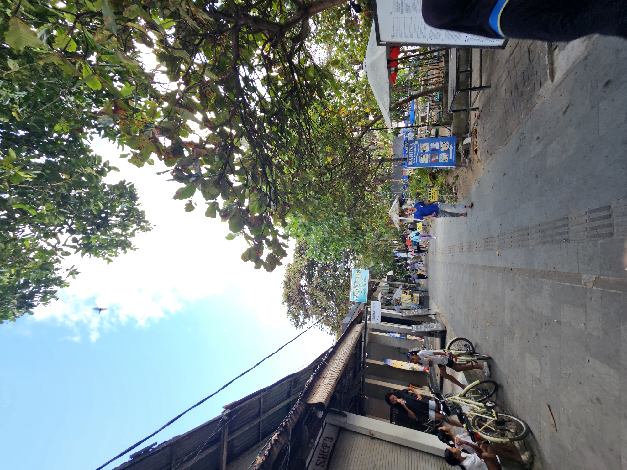 The image depicts a street scene in Denpasar, Indonesia, likely a tourist area near a beach. The foreground features a paved walkway lined with shops on the left and lush trees and more shops or establishments on the right. Several people are visible walking along the path, and a group of young women is seated near two bicycles on the left side. The shops have outdoor displays, including surfboards and signs for services like "TOOTSIE" and "BARUNA BEACH MASSAGE." The sky is blue with scattered clouds, and the sun is shining, casting shadows. The architecture of the shops suggests a tropical environment.