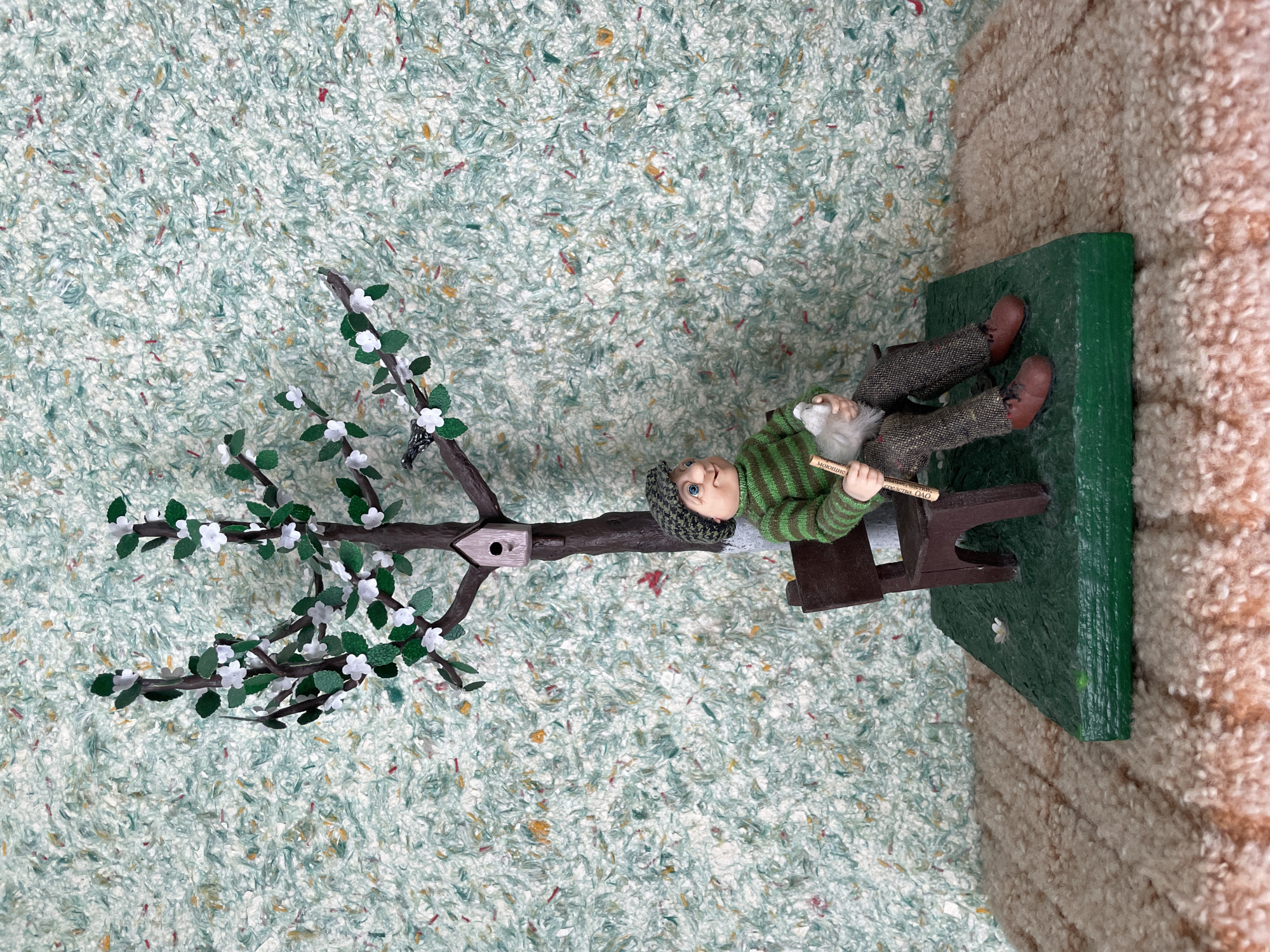 This image depicts a diorama scene of a miniature figure seated on a bench under a blossoming tree. The figure, a man wearing a green and brown striped sweater, checkered pants, a green cap, and brown shoes, is holding a white fuzzy object and a wooden stick. The tree, with brown branches, green leaves, and white blossoms, has a small birdhouse attached to its trunk. A tiny black bird is perched on one of the branches. The diorama is set on a green base, which rests on a textured, carpet-like surface. The background is a textured wallpaper in shades of light green and white with flecks of other colors. There is no discernible location in the city of Perm, Russia, depicted in this image.
