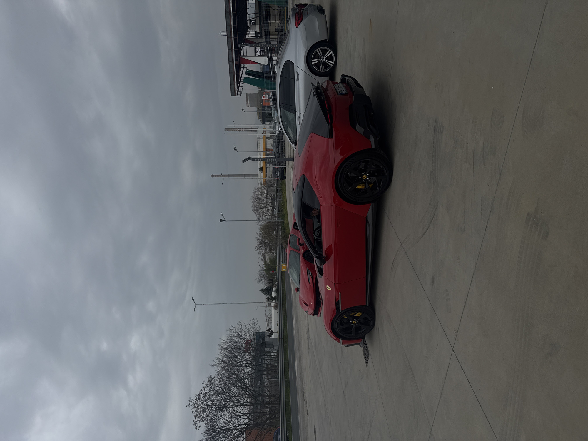 A red Ferrari, with a partially open driver-side door, is parked on a concrete surface near a road on an overcast day. A gray vehicle is parked behind it. In the background, there are buildings, trees, and utility poles, indicating an urban setting. A sign is vaguely visible near the buildings. The location is likely near the city of Plovdiv, Bulgaria.