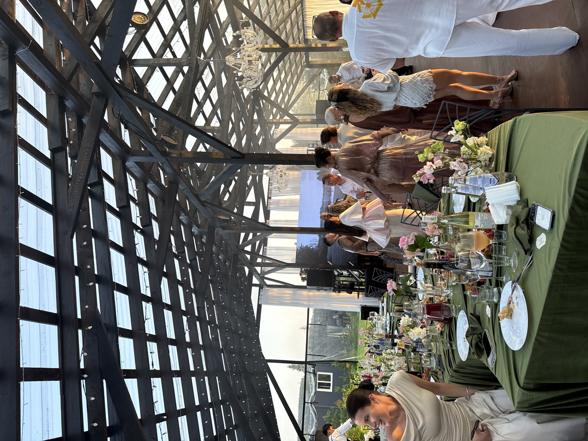 The image depicts an outdoor event under a wooden beamed structure with a transparent roof, likely in Светлое, Russia, given the user prompt. A long, green-clothed table is set for a meal with various bottles of drinks, glasses, plates, and floral centerpieces. Several people are gathered around the table and in the background, some standing and conversing, while others appear to be mingling or observing. The lighting suggests it is late afternoon or early evening, with sunlight streaming through the roof. A large screen is visible in the background, and chandeliers hang from the ceiling structure. The atmosphere appears to be festive and social.