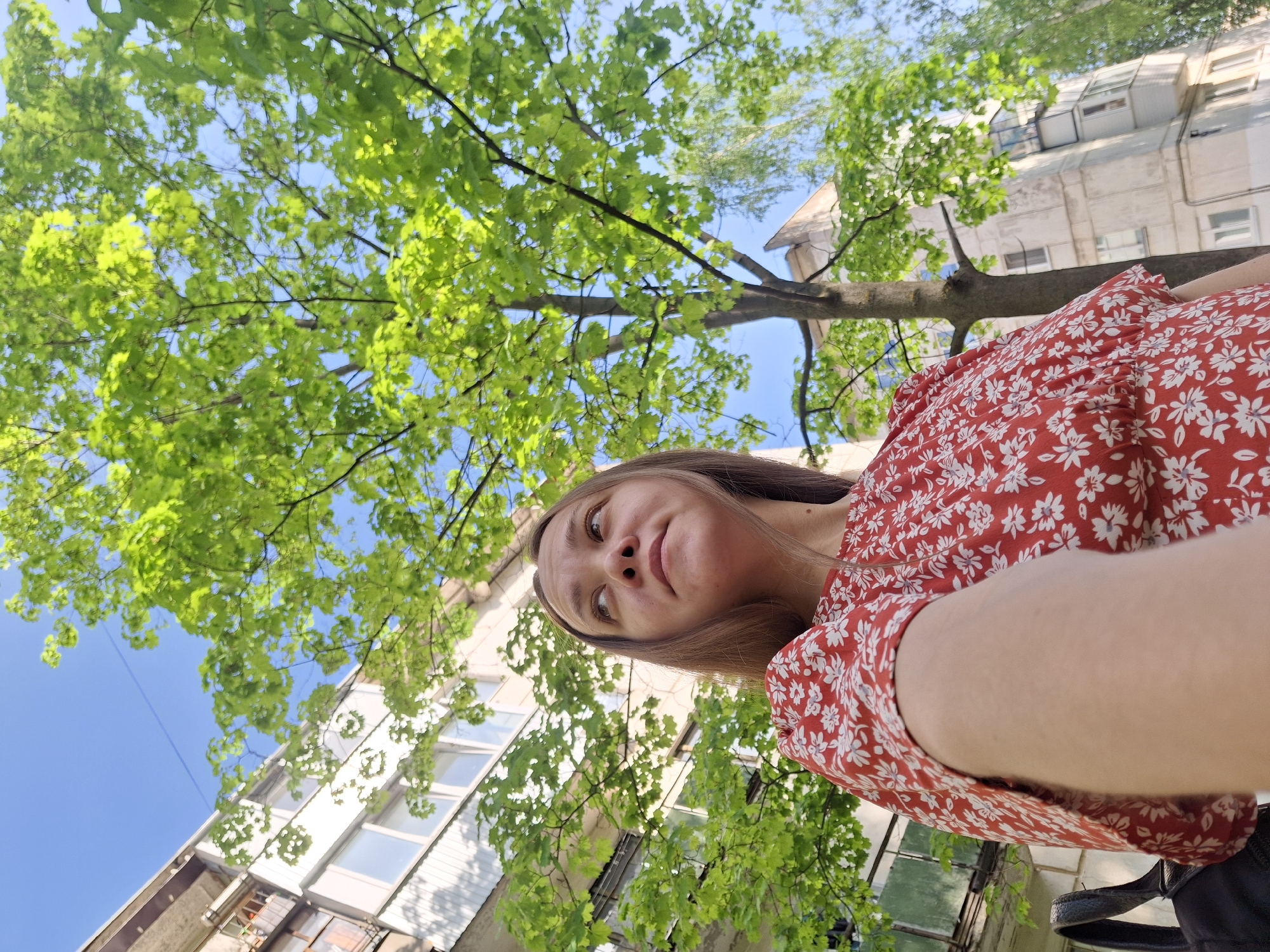 The image shows a woman in a red floral dress looking up at the camera. She is standing under a tree with green leaves, and a building is visible in the background. The sky is blue. The photo was taken in Chaikovsky, Russia.