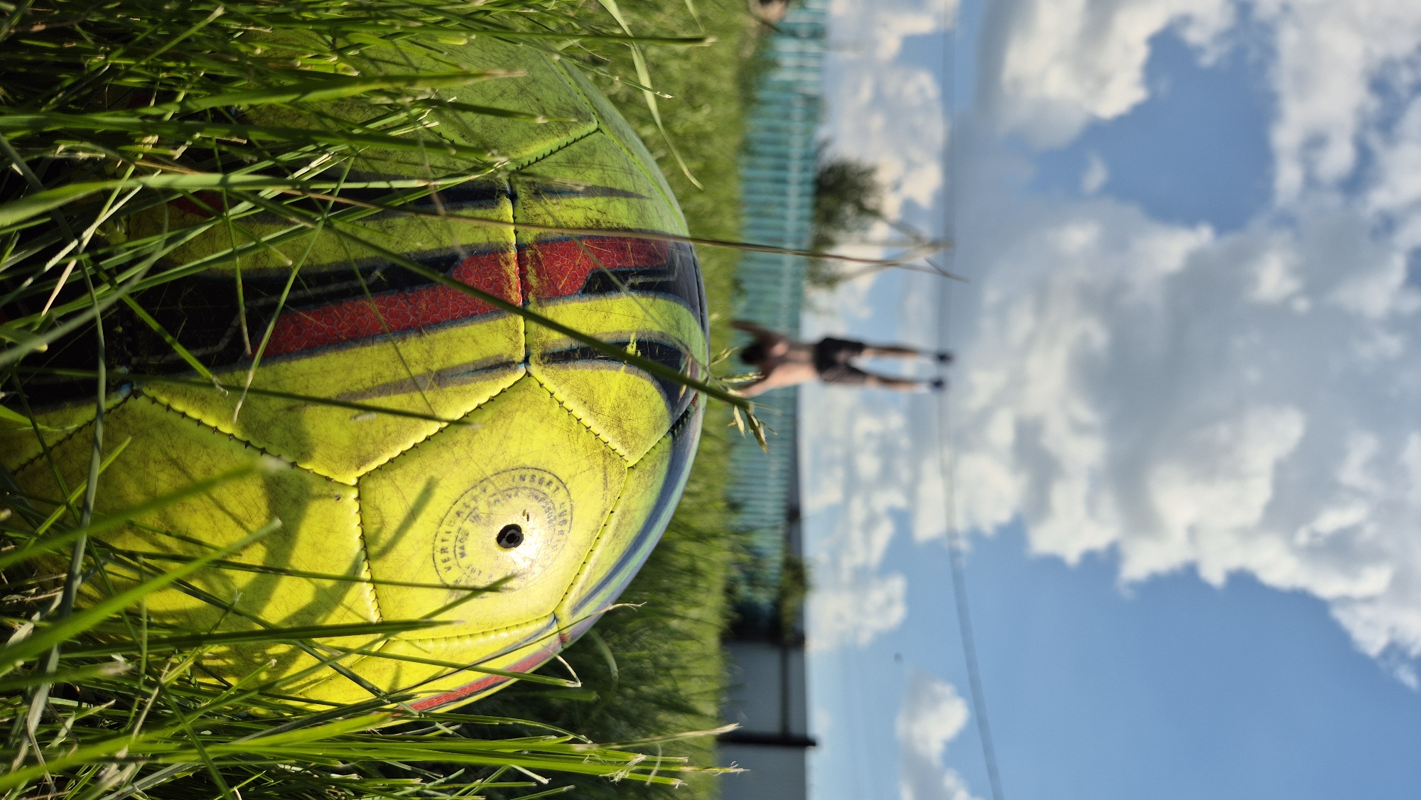 In an outdoor setting with green grass, a faded yellow and red soccer ball is prominently featured in the foreground, partially obscured by tall blades of grass. In the background, a shirtless person is performing a handstand, with their legs in the air. A light blue fence runs horizontally behind the person. The sky above is filled with white clouds against a blue backdrop. The scene appears to be in a rural or suburban area, possibly a backyard.