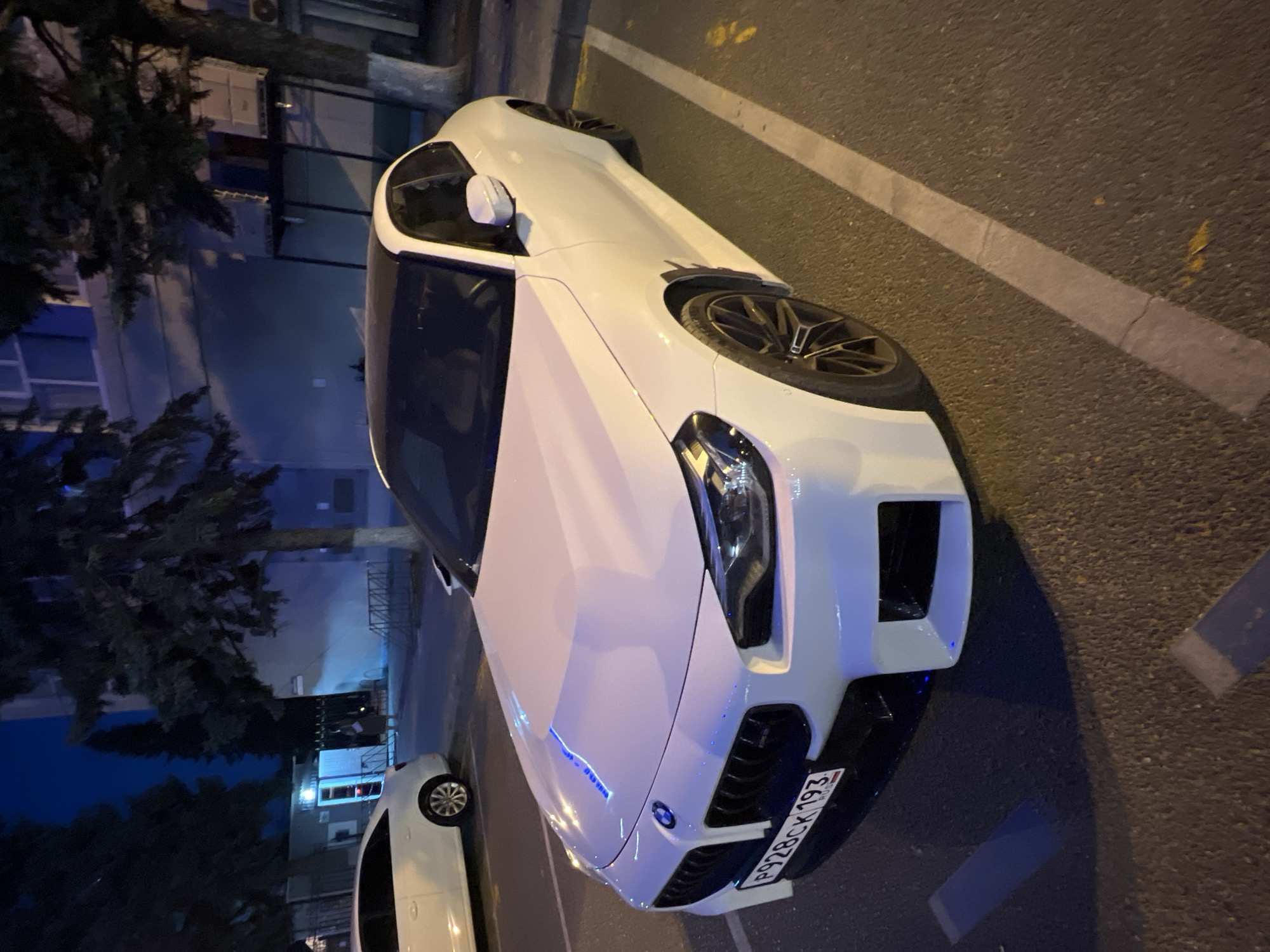 A white BMW is parked on a street at night in Krasnodar, Russia. The car is a modern sports coupe with sleek lines and prominent headlights. It has black alloy wheels and a license plate that reads "P928CK 193". The scene is dimly lit, with the car's headlights and interior lights casting a blue glow on the asphalt. The background shows a building with windows and some trees. The weather appears to be clear, and the time of day is night. There are no people visible in the image, and the overall mood is calm and still. The car is the central focus of the image, with its polished exterior reflecting the ambient light. The asphalt road is marked with white lines, indicating a parking area.