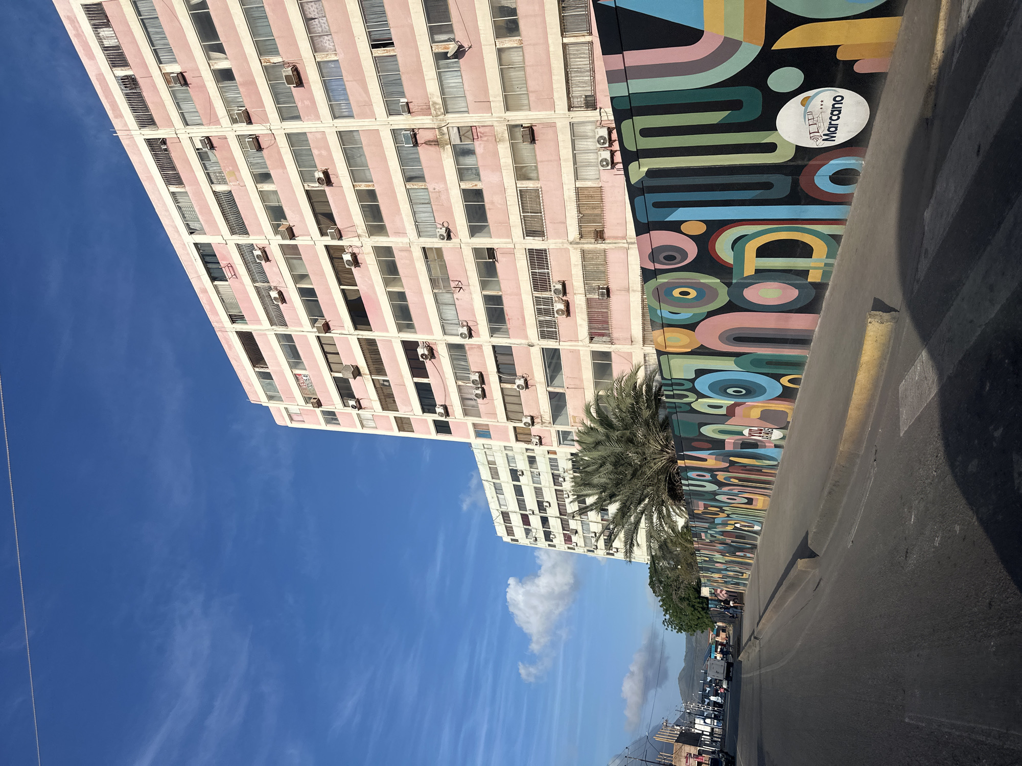 In the city of Juan Griego, Venezuela, a wide asphalt road curves to the left, bordered by a raised concrete curb and sidewalk. A long, vibrant mural depicting abstract shapes in various colors, including circles, arcs, and stripes, adorns a low wall along the sidewalk. The mural features a circular emblem with text and an image. Behind the wall and mural, palm trees and other greenery are visible, with two large, pale pink apartment buildings towering in the background under a bright blue sky with wispy white clouds. The apartment buildings have numerous windows and air conditioning units mounted on their facades. In the distance, hills are faintly visible. Several vehicles, including cars and a truck, are present on the road and sidewalk, and a few pedestrians can be seen walking along the sidewalk. The scene appears to be a street-level view of an urban environment with significant artistic and architectural features.