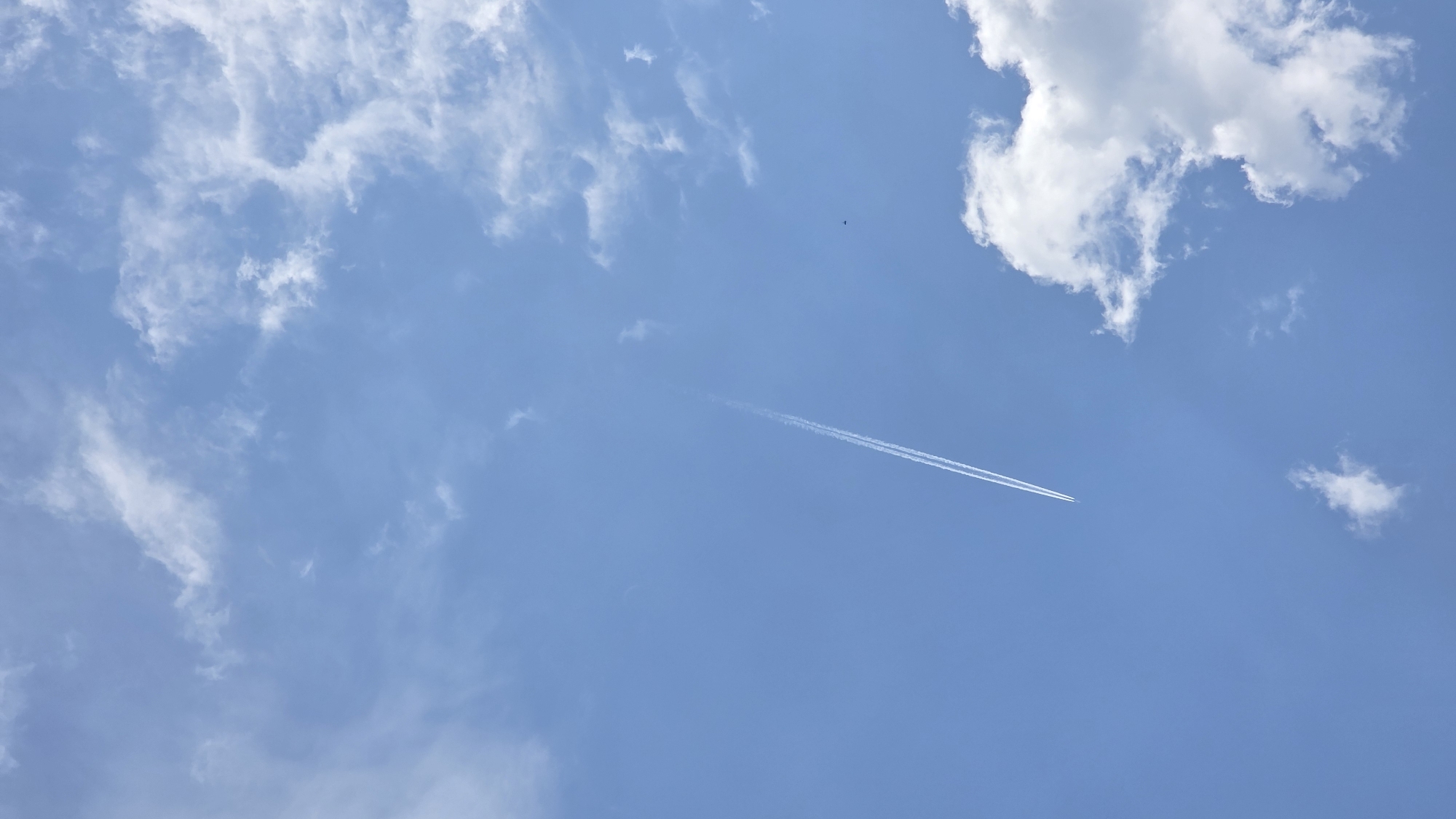 The image presents a wide-angle view of a bright, clear blue sky, partially adorned with white clouds. The sky dominates the frame, showcasing varying shades of blue indicative of a sunny daytime.
Scattered across the expanse are numerous white clouds. In the upper left quadrant, there are wispy, diffused cirrus clouds, while the upper right features a more prominent, distinct cumulus cloud formation that appears fluffy and well-defined. Smaller, dispersed cloud patches are also visible throughout the frame.
A notable element is the long, linear white contrail streaking diagonally across the sky from the lower-left to the upper-right. This contrail signifies the passage of a high-flying airplane, which itself is not clearly visible beyond the trail. Slightly above and to the left of the contrail's mid-section, a very small, dark, indistinct speck can be observed, possibly a bird or another distant object.
The scene evokes a feeling of openness and tranquility, suggesting a pleasant, fair-weather day. The absence of direct sunlight sources or distinct shadows implies it could be late morning or early afternoon. No visible text is present in the image. The setting for this serene sky view is Drenkovo, Bulgaria.
