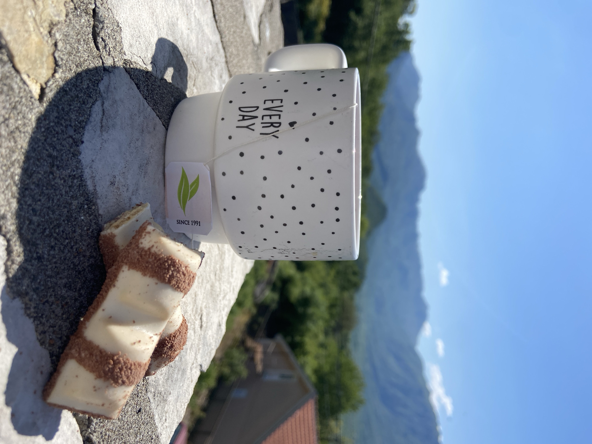 A white mug with black polka dots and the words "EVERY DAY" sits on a stone surface, accompanied by two Kinder Bueno bars. The mug has a tea bag with a green leaf logo and the text "SINCE 1991" attached to it. The backdrop includes a view of green trees, mountains, and a clear blue sky, possibly a scene within the city of Sotonići, Montenegro. A house with a red tile roof is partially visible below the trees.
