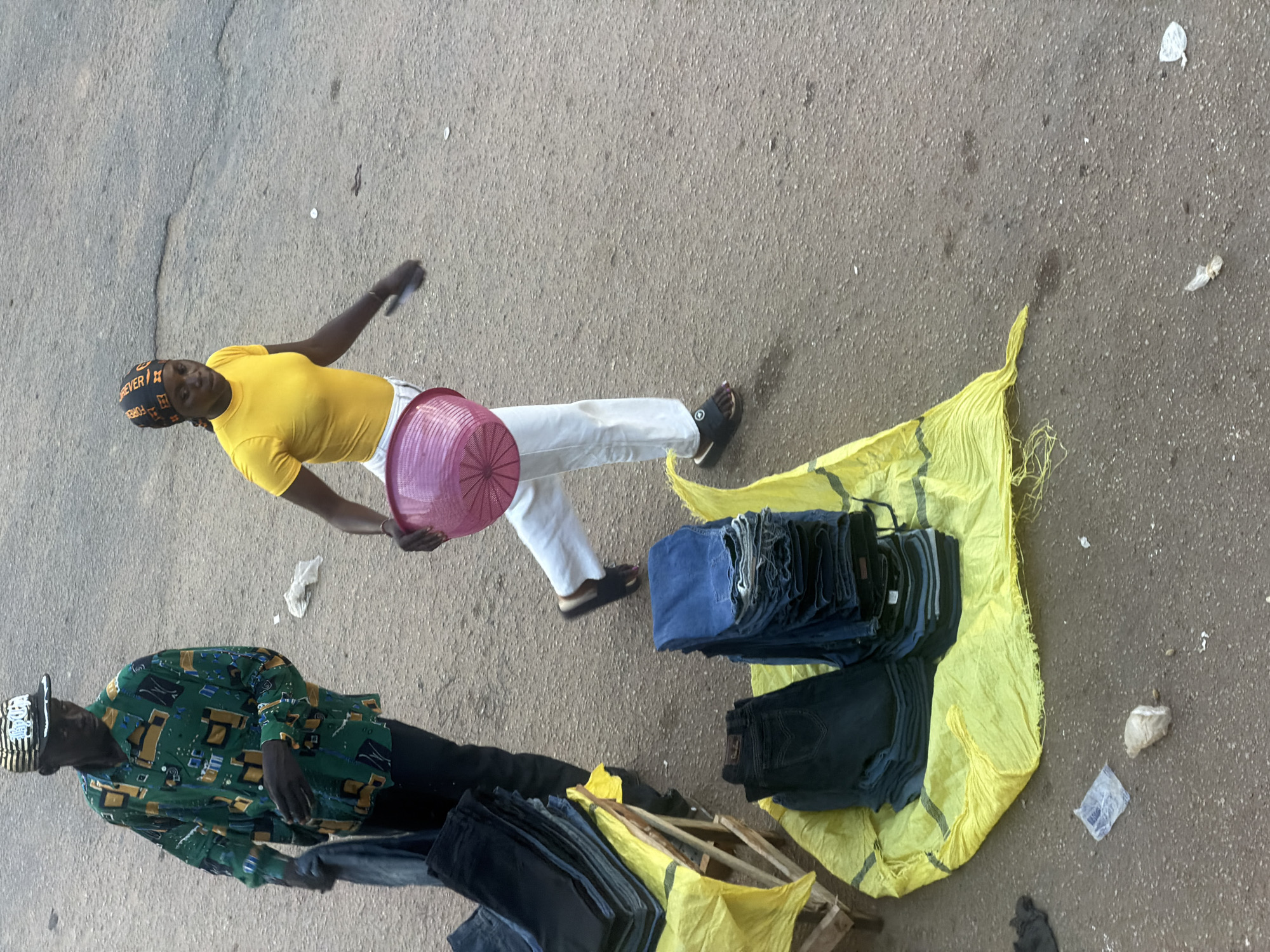 Two individuals are observed in an outdoor, urban environment, likely in Bukuru, Nigeria. The scene appears to be a street market.
On the left, a person wearing a vibrantly patterned green long-sleeved shirt and dark trousers, along with a striped baseball cap, is holding a folded dark garment in their left hand. This individual is standing near a wooden crate stacked with folded dark trousers.
To the right, a person is walking, wearing a bright yellow t-shirt, white trousers, and dark sandals. They are carrying a pink woven basket in their right arm. A stack of folded blue jeans is visible on a yellow sheet to their right.
The ground is a paved, asphalt surface with visible debris, including plastic wrappers and small scattered objects. The overall lighting suggests it is daytime.