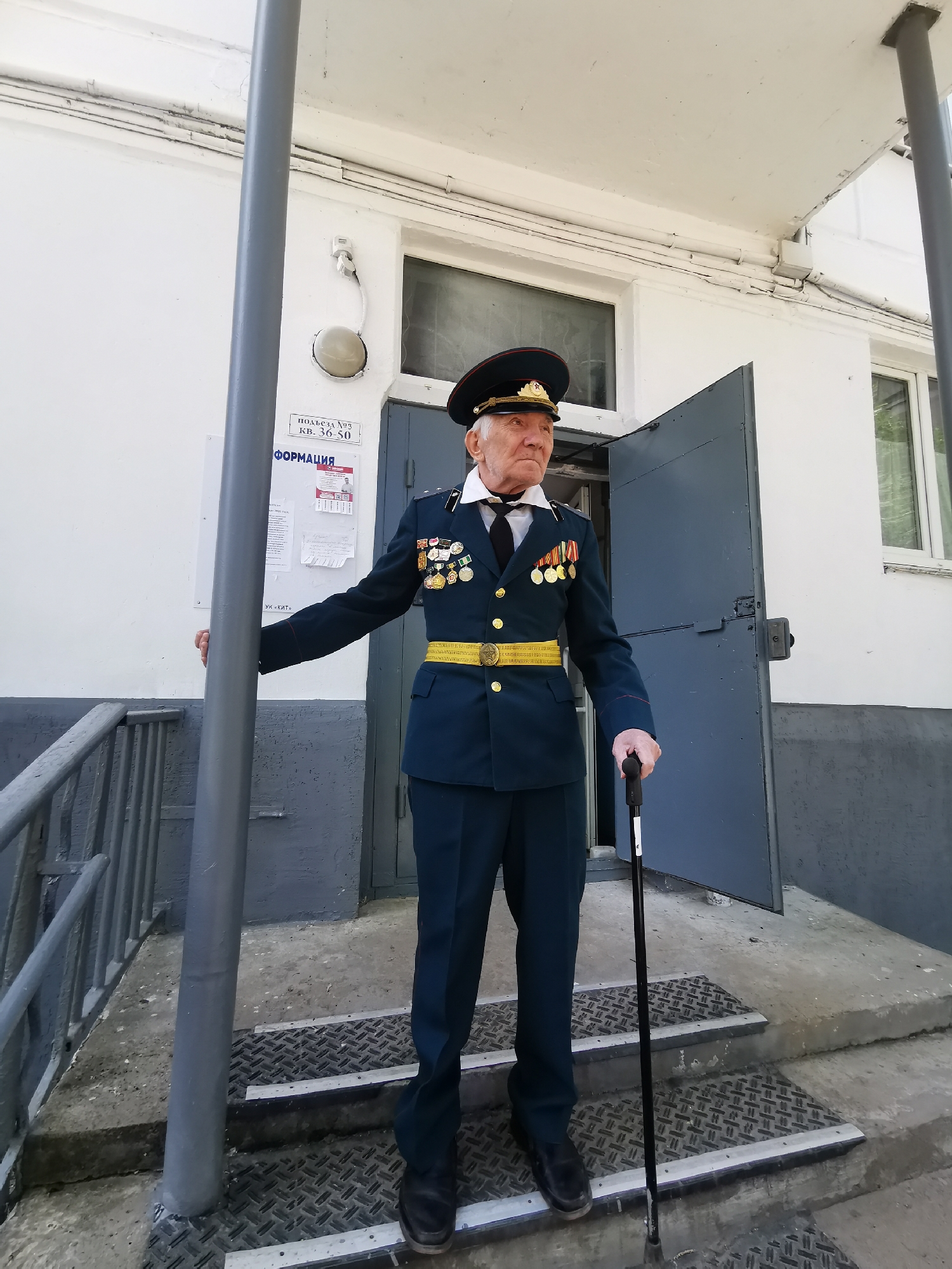 An elderly man in a dark blue military uniform stands on the steps outside a building in Chaikovsky, Russia. The uniform includes a yellow belt and numerous medals on the left breast. He wears a military cap and a white shirt with a black tie. He supports himself with a black cane in his right hand and rests his left hand on a grey metal pole. The building is painted white with grey accents and has a door slightly ajar behind him. A sign with Russian text is visible on the wall. Metal railings are present on the left side of the steps, which are covered with a metal diamond plate pattern.