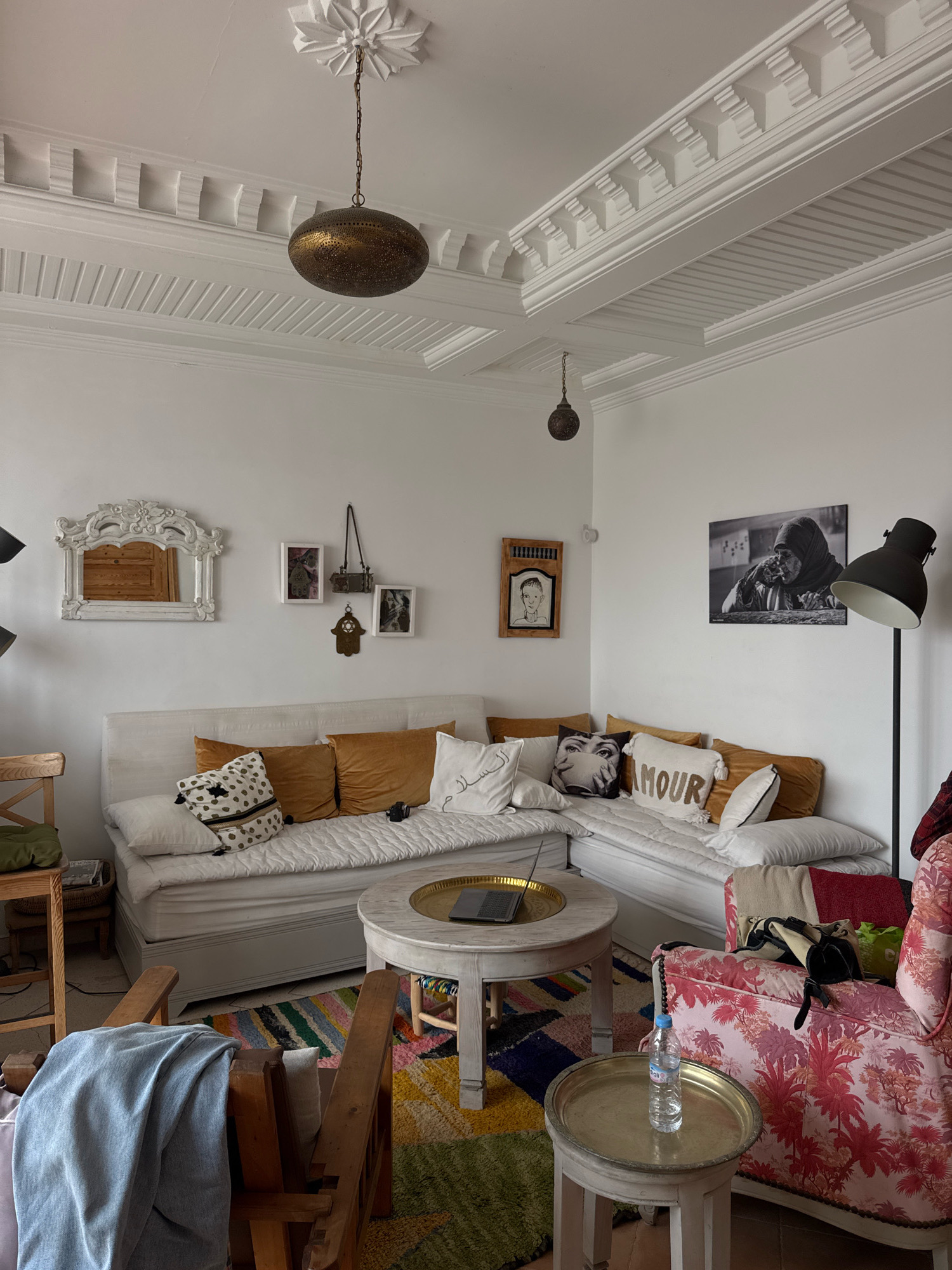 This image depicts an interior scene in a room that appears to be located in Essaouira, Morocco. The room is furnished with a large, sectional white sofa adorned with several decorative pillows in various colors and patterns, including one with the word "AMOUR" and another featuring a close-up of eyes. A round, light-colored coffee table with a golden tray sits in front of the sofa, holding a laptop. To the left of the sofa, a decorative white mirror with ornate detailing hangs on the wall above a wooden door. The walls are white and feature intricate white ceiling molding. Two distinctive pendant lights hang from the ceiling, one oval and metallic, the other spherical and darker. On the wall to the right of the sofa, a large black and white photograph of a woman wearing a headscarf is displayed. A tall, black floor lamp stands next to the photograph. In the foreground, a wooden chair is draped with a light blue blanket. A colorful, patterned rug covers the floor. Another small table with a bottle of water sits to the right of the coffee table, next to a patterned armchair. The overall impression is that of a well-decorated living space with a bohemian or eclectic style.