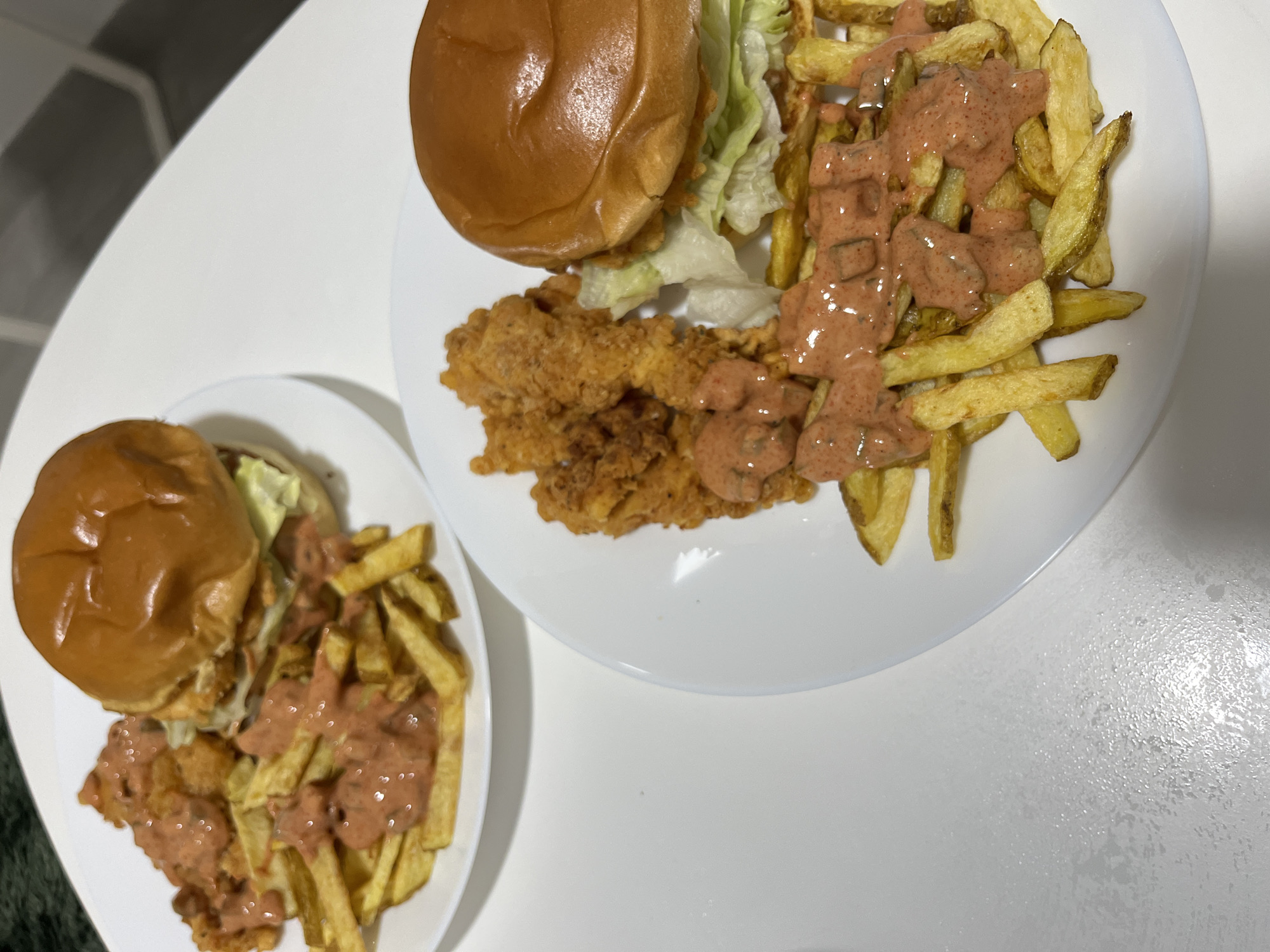 On a white surface are two white plates. Each plate contains a burger and fries with a sauce. The plates are round. The burger consists of a patty, a bun, lettuce, and sauce. The fries are topped with a pinkish-orange sauce with what appear to be chopped pickles in the sauce. There are no people visible in the image. The location of the image is not identifiable as Mogán, Spain, and there are no environmental context clues.