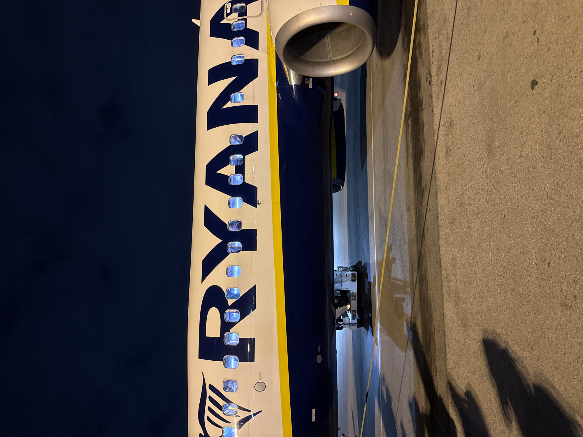 The image shows the side of a Ryanair airplane parked at an airport. The exterior is painted in the airline's signature blue and yellow colors, with the word "Ryanair" prominently displayed. The windows of the plane are visible along the fuselage, and an engine is positioned above the wing. Below the plane, ground service equipment is parked. The setting appears to be at night or in low light conditions, and the location is possibly Sofia, Bulgaria, given the presence of Ryanair, which operates flights to and from Sofia Airport. The plane is on a paved surface with yellow lines marking the airport apron.