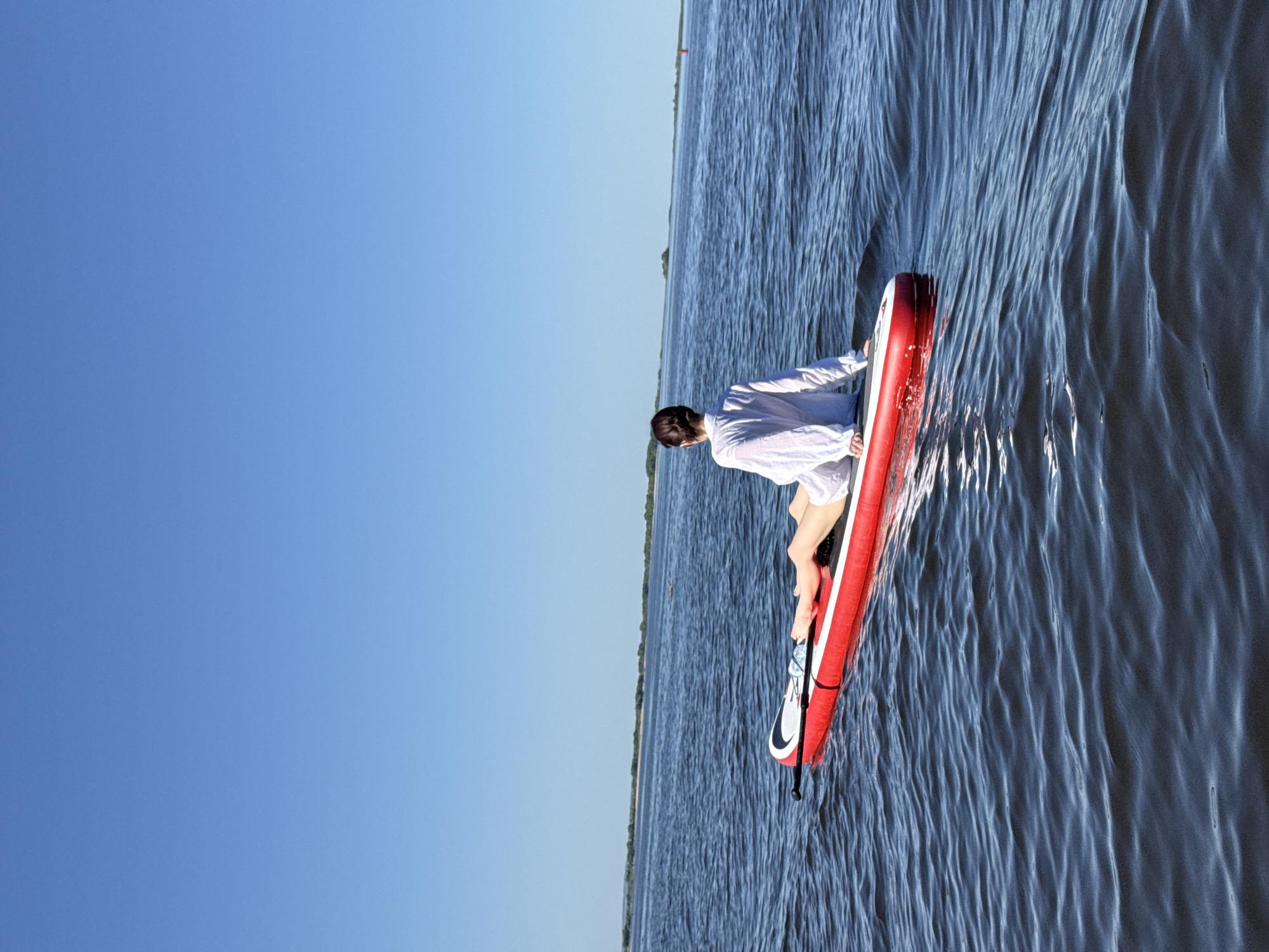 A person in a white shirt is on a red and white paddleboard in water. The water has ripples and a shoreline is visible in the distance. The sky is a clear blue. The location is near Ust'-Kurdium, Russia.