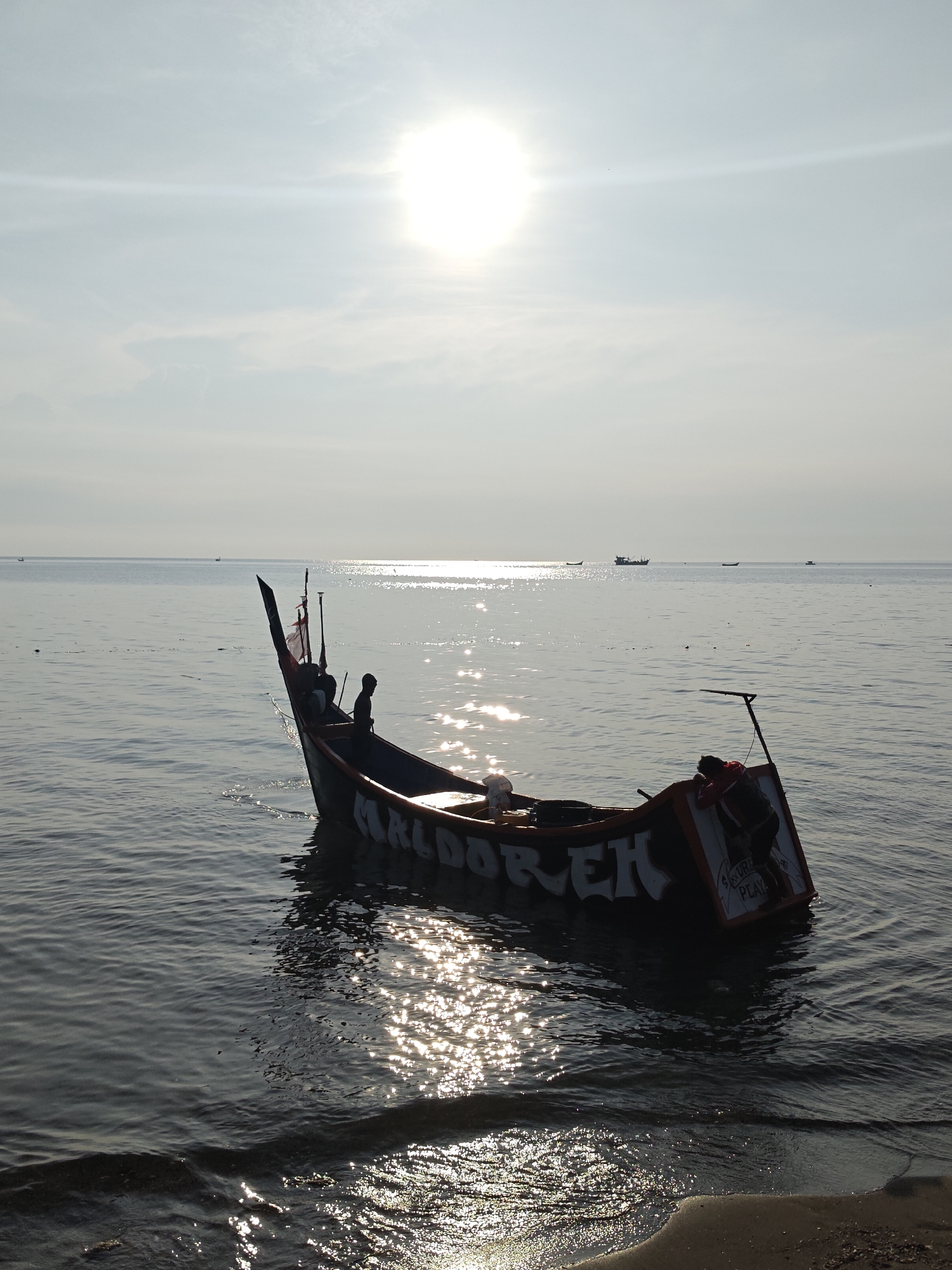 The image shows a fishing boat partially submerged in shallow water near a beach. The hull of the boat has the name "MALDO/REH" painted on it in white letters. A person is visible inside the boat. The beach appears to be located near the city of Lhokseumawe, Indonesia. The sun is high in the sky, creating glare on the water's surface. A few other boats are visible in the distance. The shoreline is sandy and gently sloping. The water is calm.
