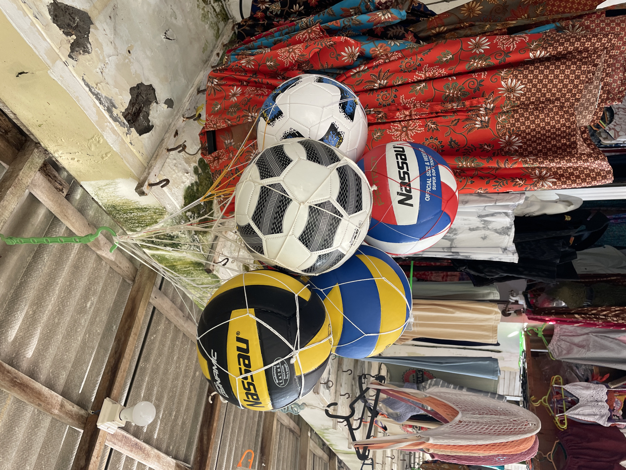 The image captures a detailed view inside what appears to be a market stall or shop, likely in Kuta Selatan, Indonesia. The central subjects are five sports balls, prominently displayed and hanging in netting. There are two black and white soccer balls, one white and blue soccer ball, one red, white, and blue volleyball, and two yellow and blue volleyballs. The red, white, and blue volleyball is branded "Nassau" with text reading "OFFICIAL SIZE & WEIGHT SUPER SOFT TOUCH." One of the yellow and blue volleyballs also features the "Nassau" brand, along with "SINAMIC" and "Manufactured LEATHER REGULATION Size & Weight ALL CONDI".
Behind the hanging balls, neatly folded and stacked textiles are visible, featuring rich red, blue, and brown fabrics adorned with intricate white, orange, and brown floral and paisley patterns, possibly indicative of batik or similar traditional prints.
The ceiling and upper structure of the building show signs of age and wear. Portions of the white ceiling plaster are peeling away, revealing darker material beneath, and there are visible patches of green mold or mildew. A corrugated metal roof is seen in the upper left, supported by rustic wooden beams. An exposed light bulb in a white ceramic socket is attached to one of these beams, providing illumination.
To the right and in the lower background, more merchandise is visible, including various clothes hanging on racks and other items stored, suggesting a wider array of goods for sale. A white mesh bag or net is also partially visible among these items. Another piece of text, "TANG", is partially visible on an item in the lower right. The overall scene suggests a commercial setting for selling goods, with no people visible. The specific time of day is not evident, given the indoor setting and artificial light source.