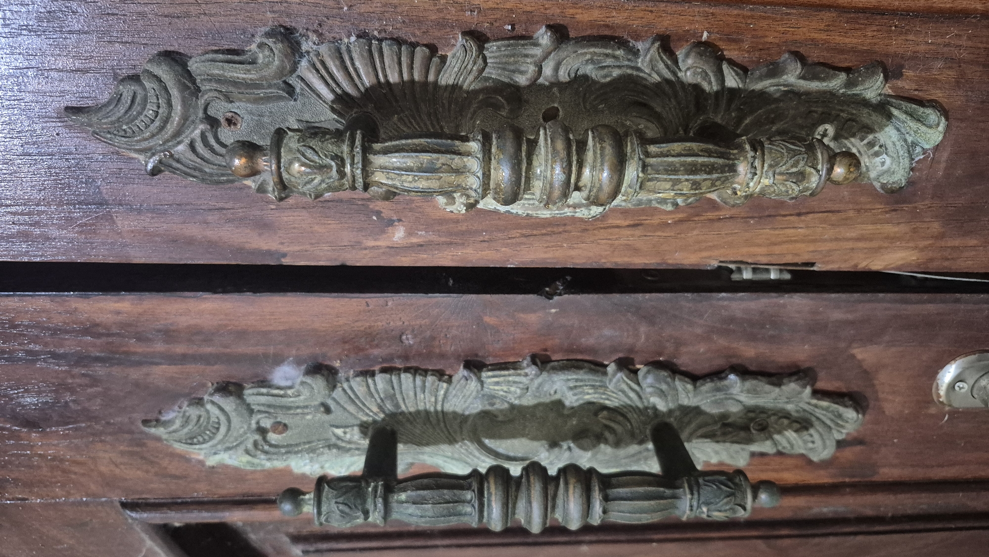 The image displays two ornate, antique brass door handles mounted on a wooden door. The handles are elaborate, with carved details and a weathered patina. The door appears to be made of dark wood and is slightly ajar, revealing a glimpse of another door panel and a hinge mechanism. The scene suggests an older, possibly historical, building. Without further information or context, it is not possible to determine the specific location in Bojonegoro, Indonesia. The image focuses solely on the door hardware and the adjacent wooden surfaces.