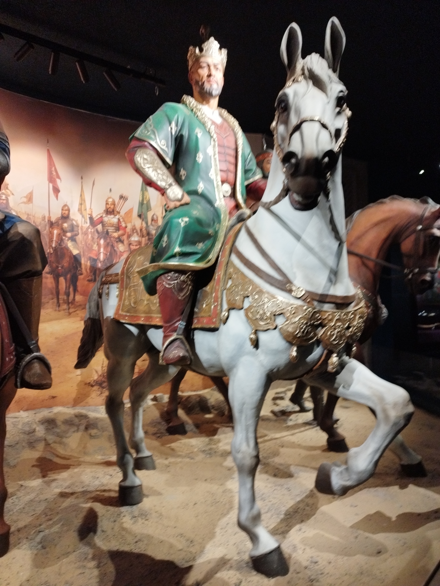 The image shows a diorama of a historical scene, likely in a museum. In the foreground, a statue of a man wearing a crown and a green and gold robe is mounted on a white horse. The horse is adorned with decorative golden armor. The man has a beard and is looking forward. To the left of the main figure, part of another statue of a person on a horse is visible. In the background, a painted mural depicts a battle scene with many figures on horseback carrying flags and weapons. The ground in the foreground appears to be sand or dirt. The lighting suggests an indoor exhibition space. The city and specific location are unknown.