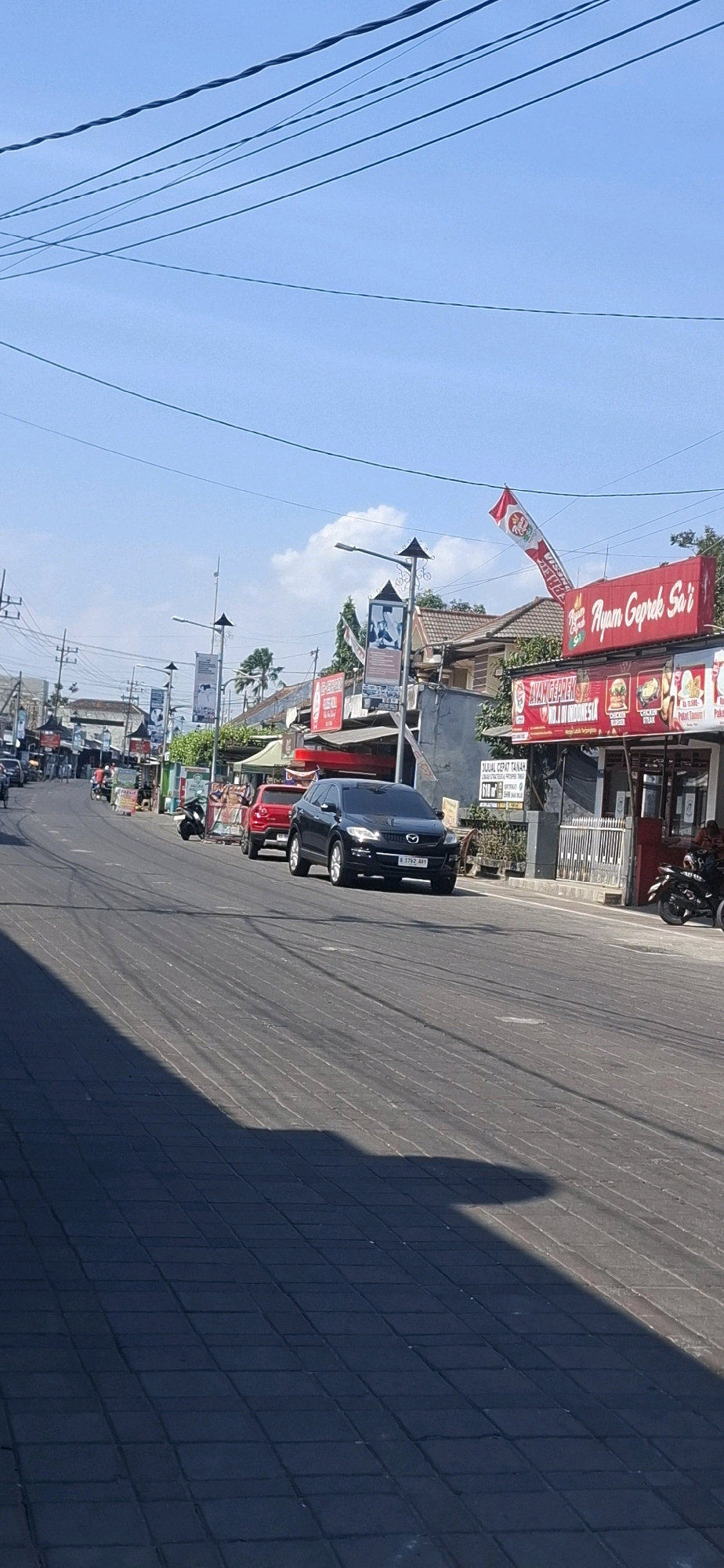 The image depicts a street scene in Blitar, Indonesia, on a clear, sunny day. A paved road occupies the foreground, with a distinct shadow cast across it from an unseen object on the left. The road curves slightly to the left as it recedes into the distance, lined with buildings and shops on the right side of the frame.
Several vehicles are present on the road. A black SUV, identified by a license plate starting with "B 1762 AAY," is stopped in the right lane. To its left, a red SUV is also stopped. Further down the street, a few motorcycles are visible, along with other cars and people.
The buildings lining the street include commercial establishments with signage. Notably, a red sign with white lettering reads "Ayam Geprek Sai," indicating a food establishment. Another sign below it advertises "AYAM GEPREK IN INDONESIA." Other banners and signs are displayed on poles along the street. Power lines crisscross the sky, which is a bright, clear blue. Palm trees are visible behind some of the buildings. The overall impression is of a typical day on a busy urban street in Indonesia.