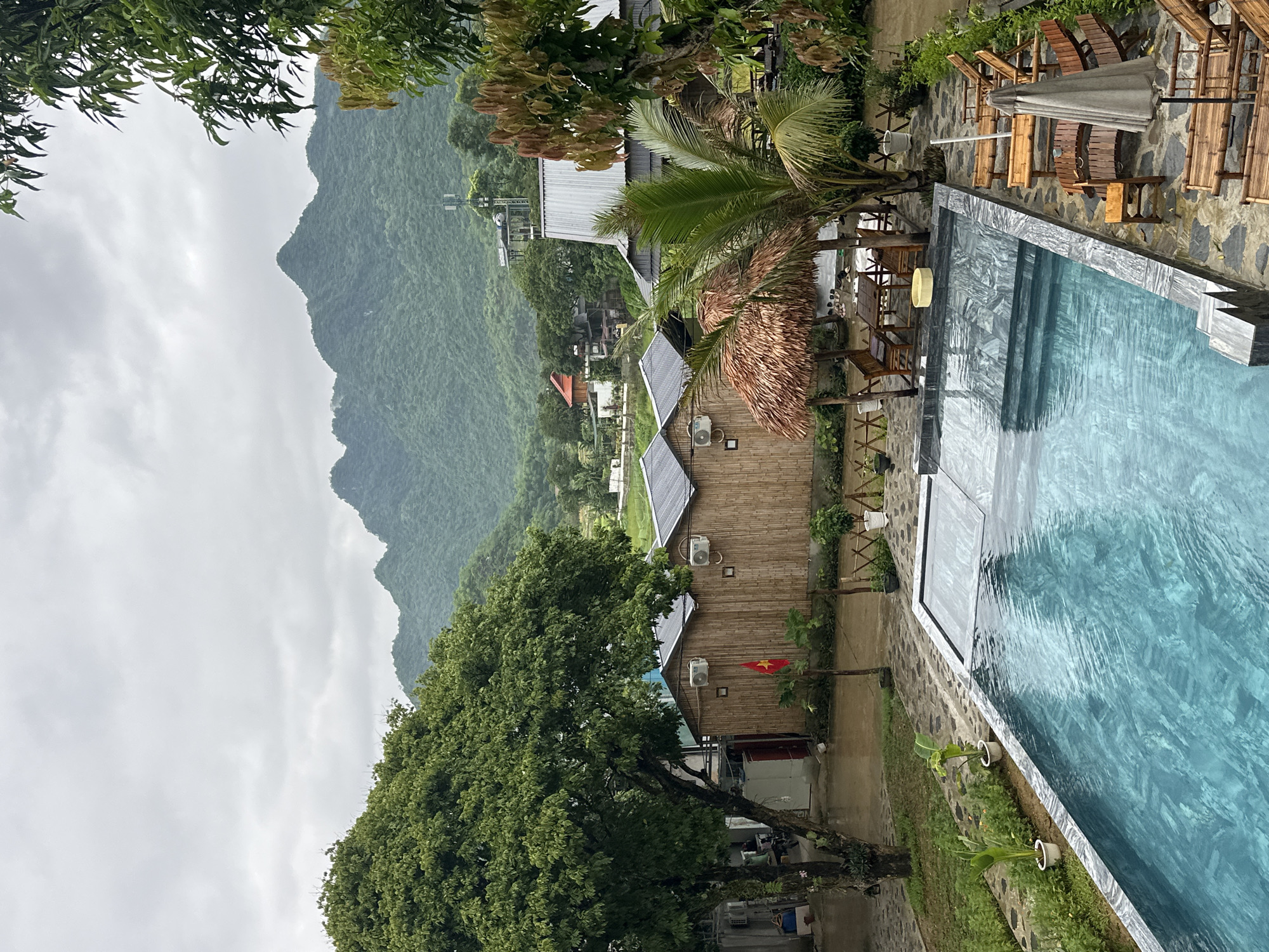 A wide-angle, eye-level shot captures a resort scene in Xóm Khán, Vietnam. The foreground features a swimming pool with turquoise water, bordered by stone tiling. A row of wooden lounge chairs with cushions and a closed umbrella are arranged on a paved area beside the pool. Behind the pool, several thatched-roof huts constructed from bamboo are visible, each equipped with air conditioning units. A Vietnamese flag flies in front of one of the huts. Lush green trees and foliage surround the buildings and pool area. In the background, rolling green hills and mountains dominate the landscape under a cloudy sky. A cell tower is visible on one of the hills. The overall impression is a serene, tropical retreat.