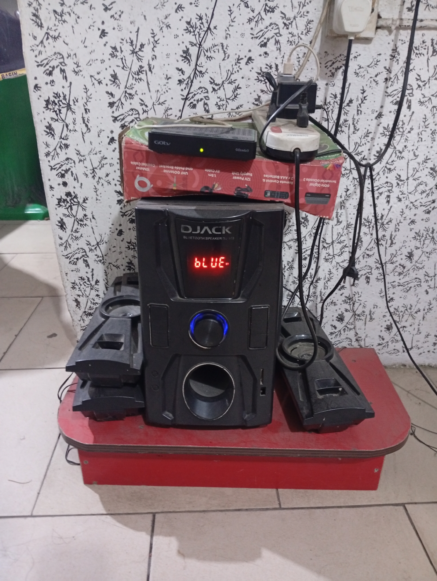 In an unidentified part of Lokoja, Nigeria, a black Djack Bluetooth speaker is centrally placed on a red wooden table. Stacked on the speaker is a Goly digital TV receiver box. An electrical outlet with multiple plugs and chargers is visible on the patterned wall behind the setup. Speaker components are scattered on the table around the main unit.