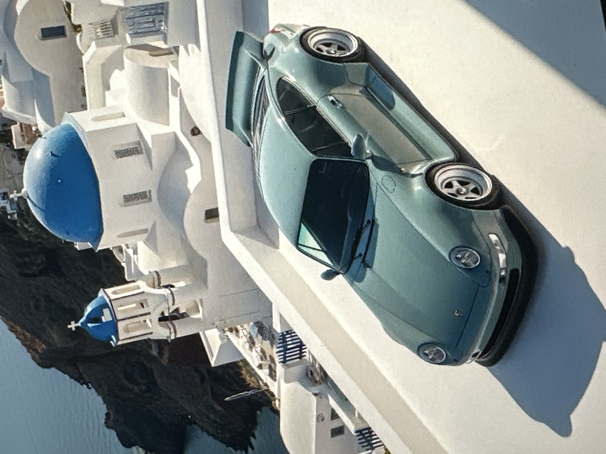 The image shows a teal-colored Porsche 911 with a prominent rear spoiler parked on a white terrace. The car is angled towards the viewer's left, with its front end slightly lower than its rear. In the background, there are white buildings with blue-domed roofs, characteristic of Greek island architecture. A steep, dark cliff face plunges down to the sea on the left side of the frame. The overall scene suggests a luxurious and picturesque setting. There is no indication that this location is in Tallinn, Estonia.