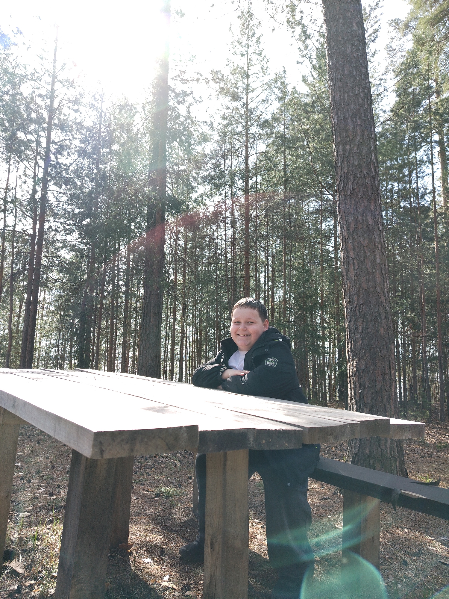 A young person is sitting at a wooden picnic table in a forest. The person is wearing a black jacket over a white shirt and dark pants. The forest consists of tall, slender trees, with sunlight filtering through the branches. The location is the city of Noviy, Russia.