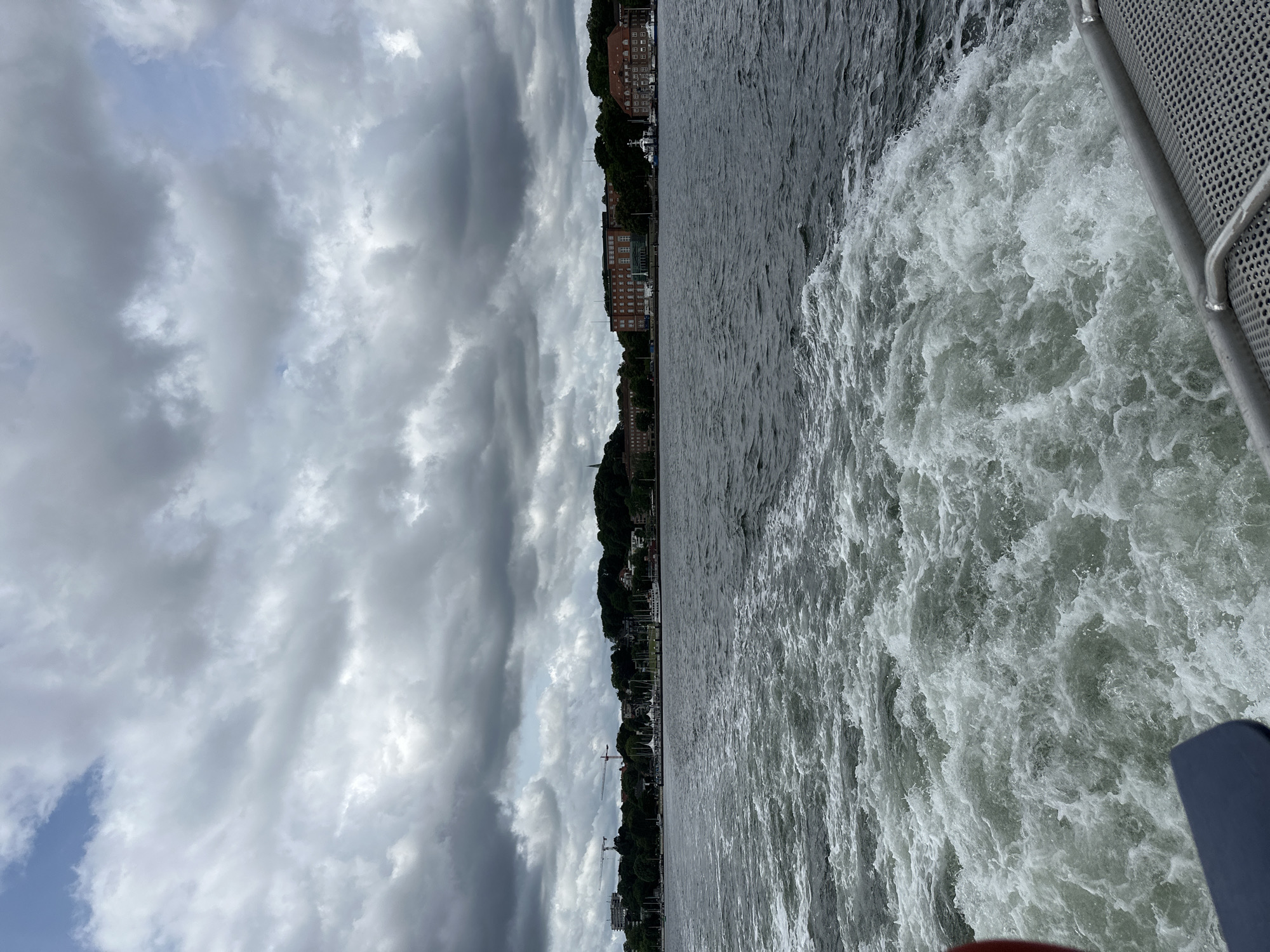 The image shows a view from a moving boat on the Kiel Fjord in Germany. The water is choppy and creating a wake behind the boat. In the distance, a city skyline is visible, with buildings and trees lining the shore. The sky is cloudy and overcast. There are no people visible in the image. The overall scene suggests a boat trip on a slightly gloomy day in Kiel.
