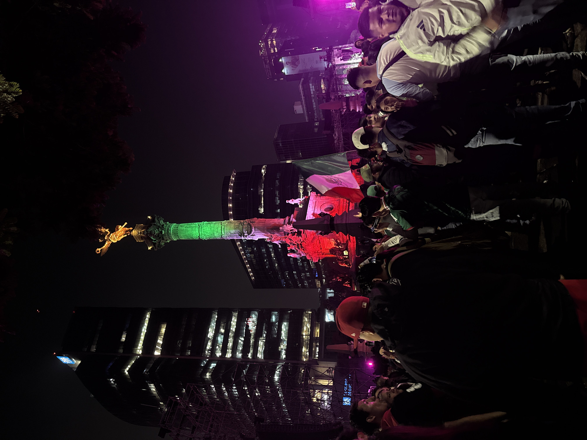 The image depicts a nighttime scene at the Angel of Independence monument in Mexico City, Mexico, during a gathering of people. The iconic monument is illuminated with green and red lights, and the golden statue of Nike at its apex is visible. A Mexican flag is prominently displayed in the foreground, held by an unseen individual. A crowd of people, many wearing casual clothing and some with hats, are gathered around the monument, suggesting a celebration or event. Tall buildings, some with illuminated windows, surround the plaza, indicating an urban setting. The atmosphere appears festive, with purple lighting in the background contributing to the ambiance. The image captures a moment of public assembly in a well-known landmark of Mexico City.