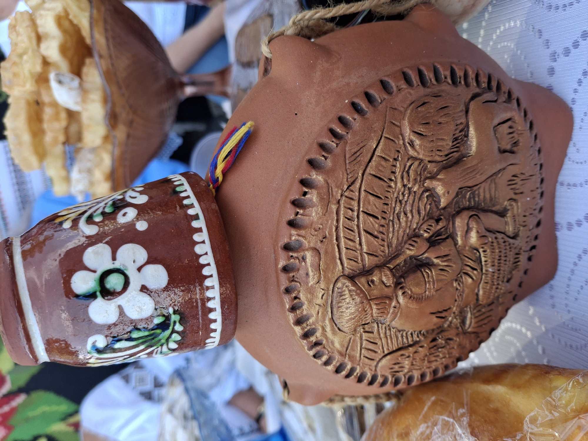 This is a close-up, eye-level shot of traditional Moldovan pottery on a white tablecloth. The pottery is reddish-brown and features intricate embossed designs. The largest piece is a flask-like object with a circular relief depicting a person with a hat, a dog, and a basket. A smaller, cap-like piece, adorned with white and green floral patterns, is placed upside down on top of the larger piece. A colorful woven string, in red, yellow, and blue, is attached to the smaller piece. In the background, a bowl filled with golden, irregularly shaped pastries is visible, along with blurred figures of people and a glimpse of a patterned textile. A loaf of bread, wrapped in plastic, is partially visible on the left. The lighting is natural and the focus is sharp on the pottery. The scene appears to be set outdoors, possibly at a market or festival in Roghi, Moldova.