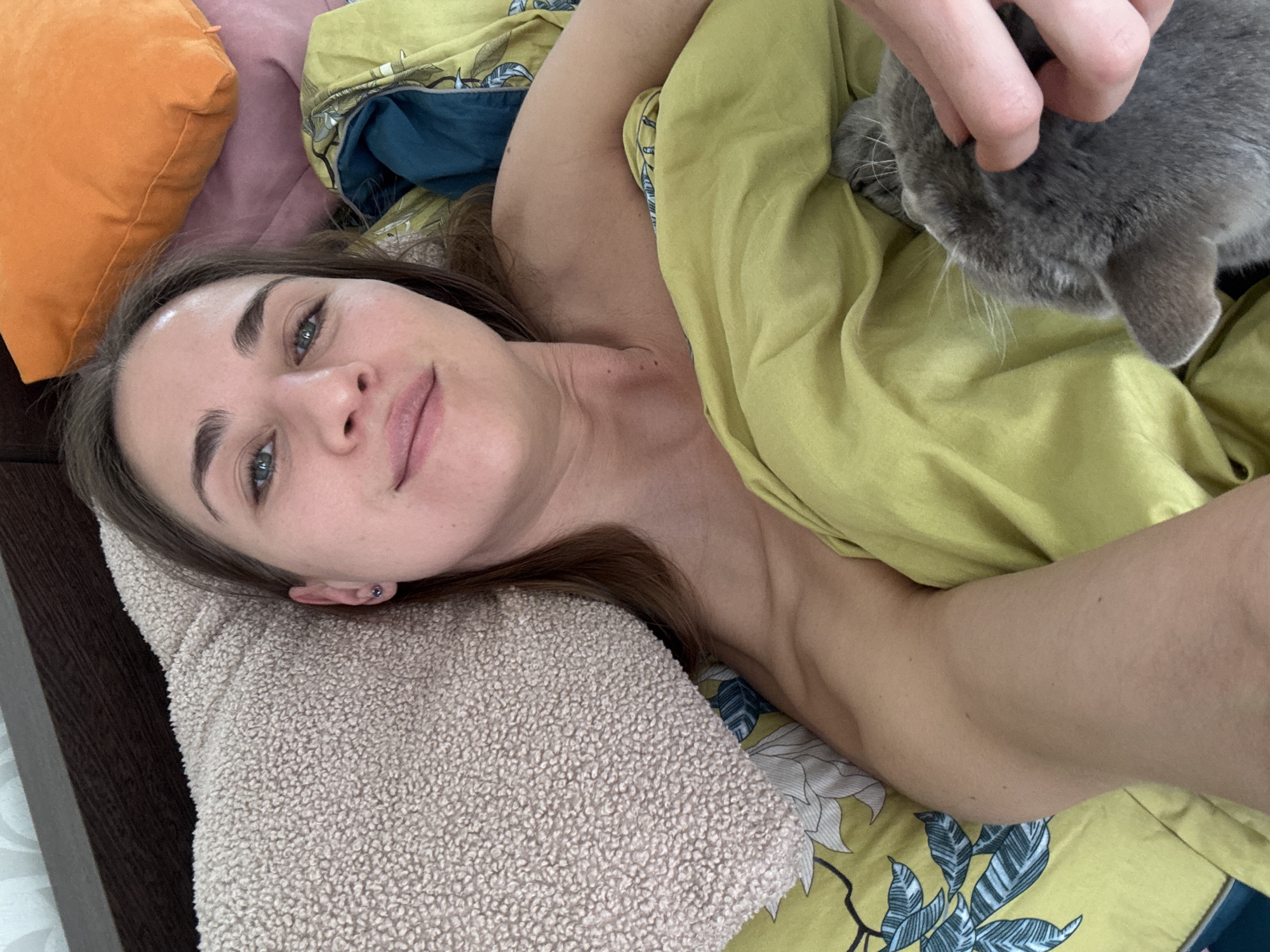 A woman is lying in bed, petting a gray cat. She is resting her head on a beige pillow next to a dark brown headboard. To her left is an orange pillow, and she is under a yellow blanket with a floral pattern. A hand is visible, petting the cat on its head. The specific location within Saint Petersburg, Russia, cannot be determined from the image.