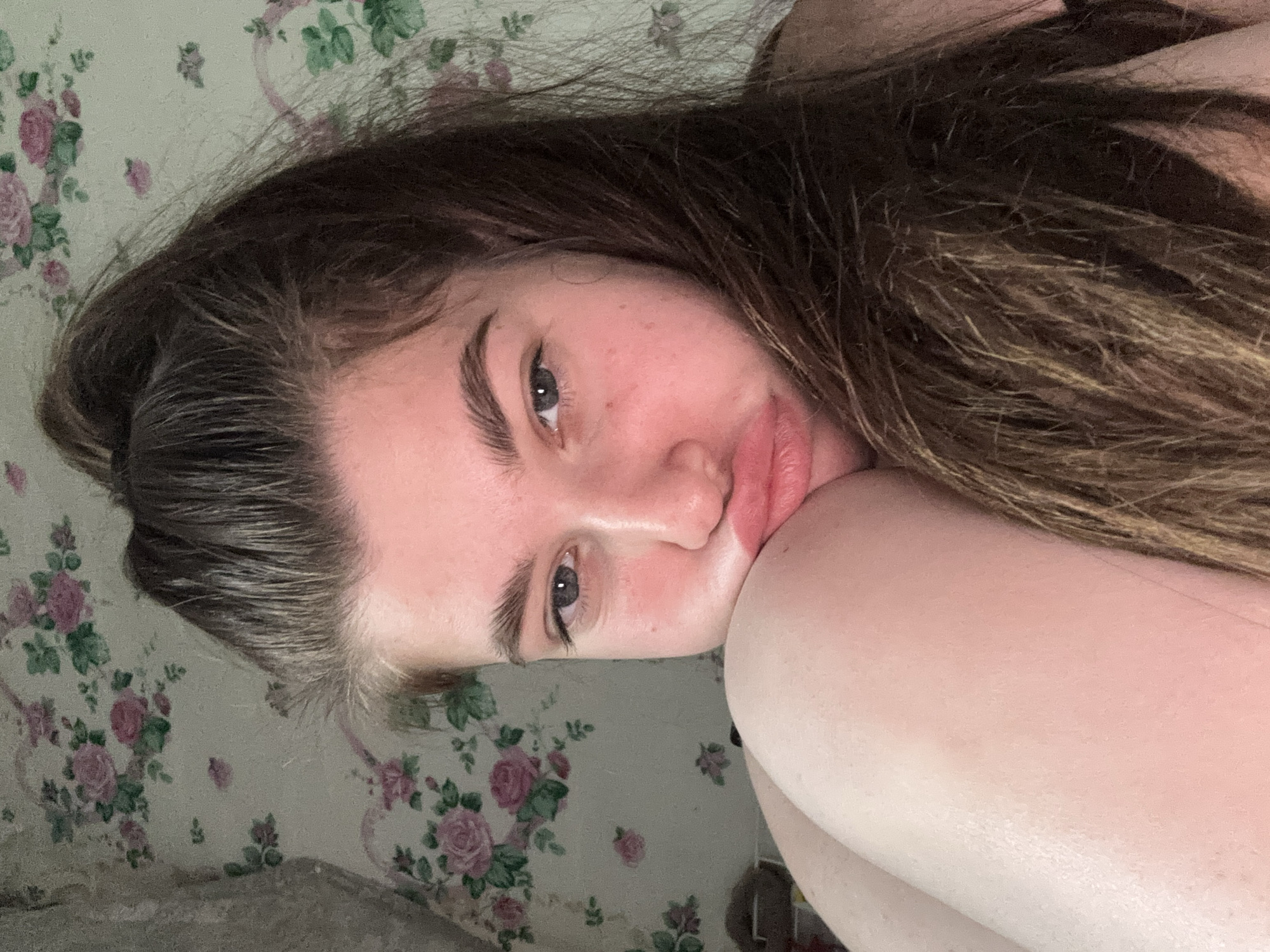 A young woman with light skin, long brown hair, and light eyes rests her head on her arm. Her facial expression appears neutral or slightly pensive. The background consists of a floral patterned wall covering or fabric with a repeated design of pink roses and green foliage.
The provided location (Мещёры, Russia) seems irrelevant to the content of the image and cannot be confirmed or related to any elements present in the scene.
