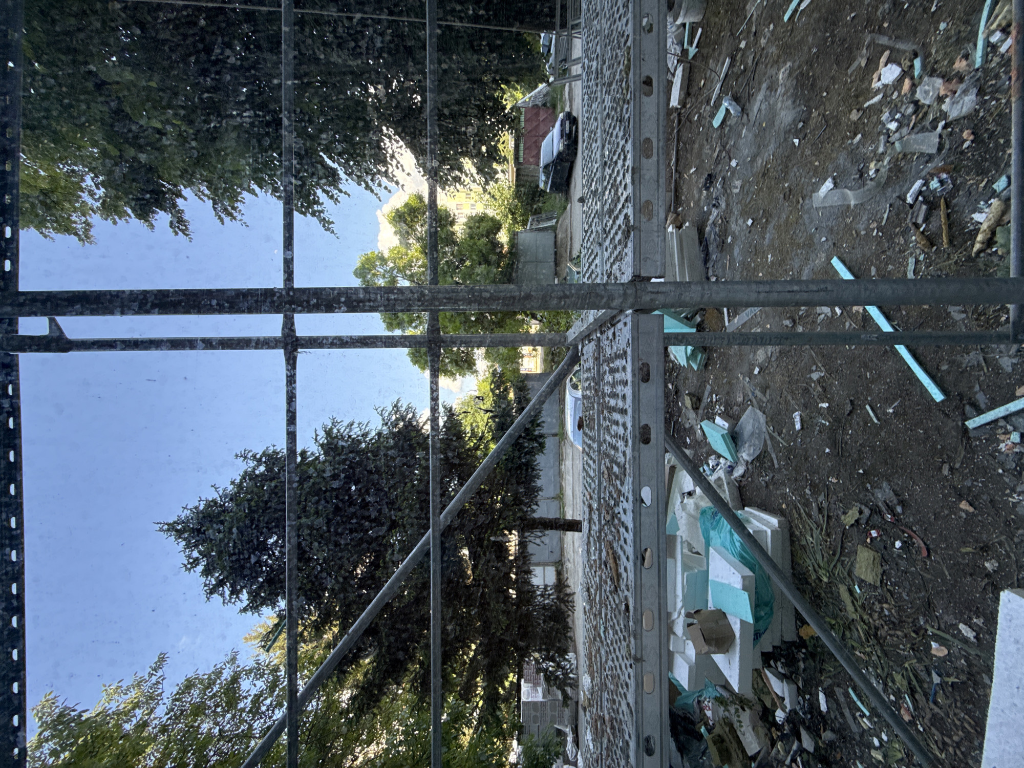 The image depicts a view from scaffolding, likely in Lovech, Bulgaria. Through the scaffolding, trees and the sky are visible. To the right of the scaffolding is a debris-strewn ground covered with construction materials and waste. Debris on the ground seems to include pieces of foam, wood, plastic, and other unidentified objects. A car and a fence are visible behind the trees in the background.