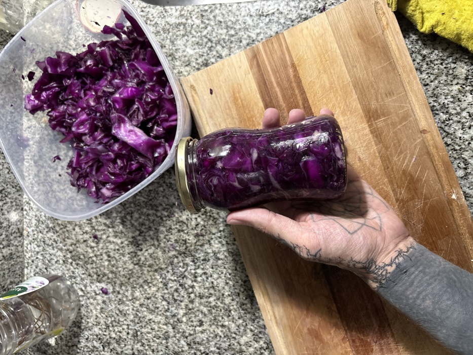 Chopped red cabbage fills a clear, square plastic container and a uniquely shaped glass jar with a golden lid. The jar is held by a tattooed hand over a wooden cutting board. The cabbage appears moist and glistening. Part of another jar sits at the bottom left of the scene. The background consists of a granite-like countertop with a yellow cloth in the top-right corner.