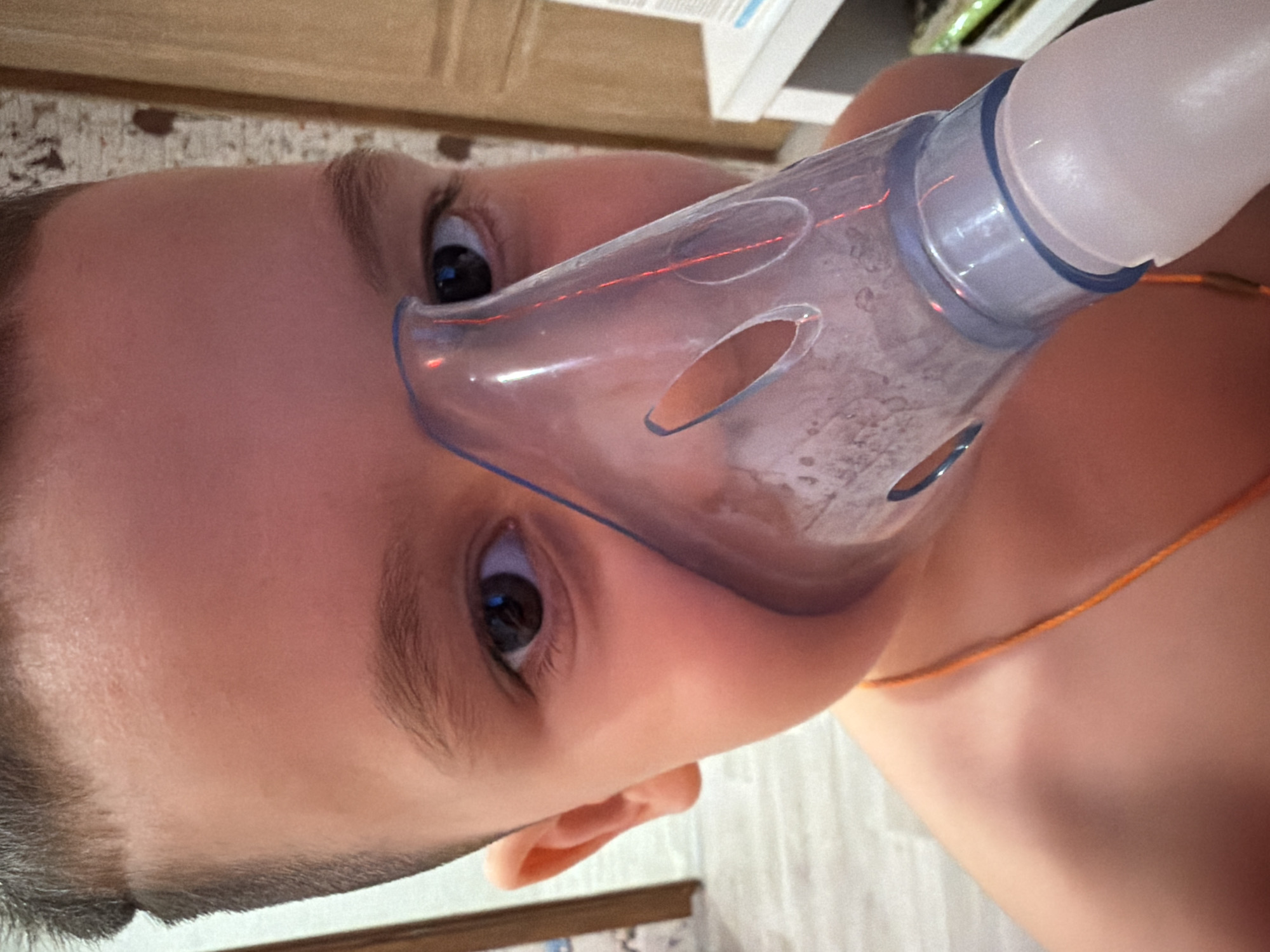 A close-up, face-forward shot shows a child wearing a nebulizer mask. The mask is made of transparent plastic with several oval holes and is attached to a white tube. The child has brown hair and dark eyes. An orange string is visible around the child's neck. The background is out of focus, but appears to be a room with white furniture. The city of Chaikovsky, Russia is not discernable from the image.