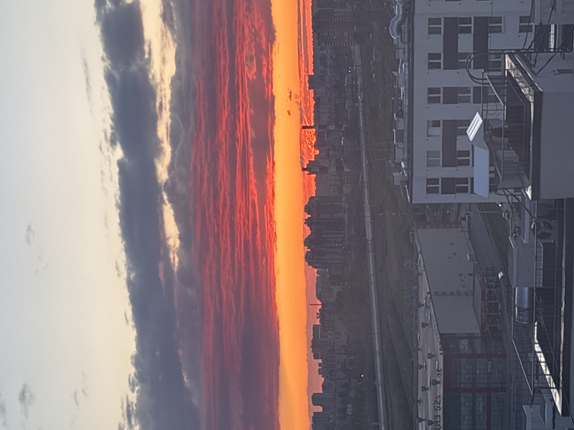 The image shows a sunset in Vsevolozhsk, Russia. A line of clouds transitions from gray to deep red. Below is a yellow-orange band, likely a sunset glow. The cityscape is silhouetted. To the right is a white building with a ladder and various utility attachments. The buildings and city elements suggest an urban environment.