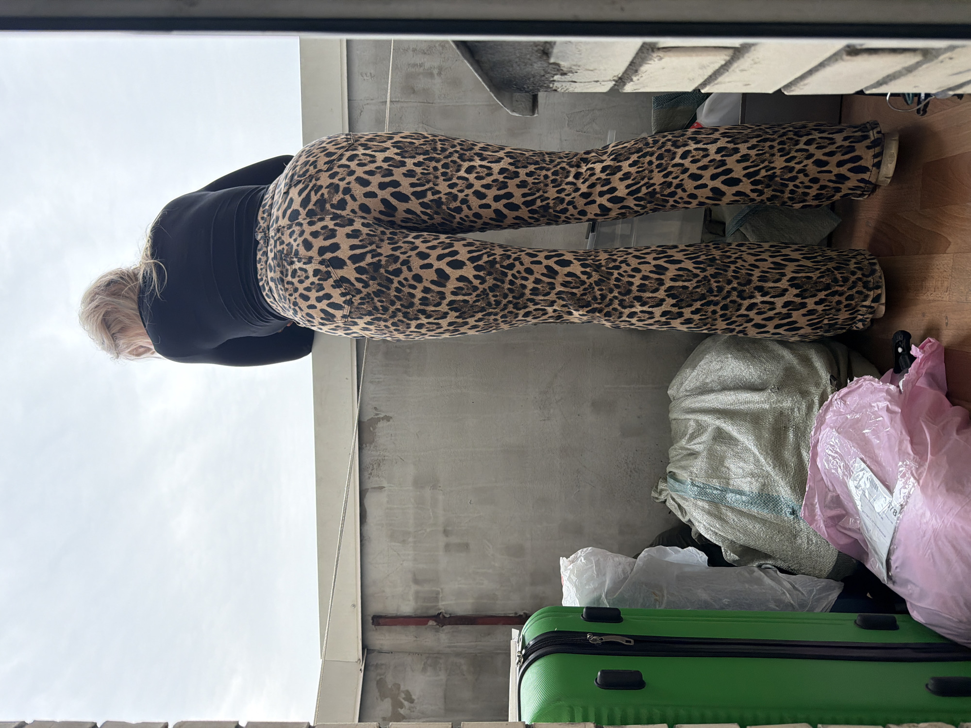 The image displays a rear view of a blonde-haired individual, presumed female, standing on a balcony or rooftop located in Saint Petersburg, Russia. The individual wears a black long-sleeved top and brown and black leopard-print flared trousers, leaning forward over a light-colored railing and looking out towards an overcast sky. To the immediate left, a bright green hard-shell suitcase stands vertically, partially obscured by the frame. Adjacent to the suitcase and extending to the background are various stored items, including a large white plastic bag, a grey fabric sack, and a pink plastic bag on the wooden floor with a white label visible on its surface. A vertical, reddish-brown pipe runs along the left concrete wall. A thin white rope or cable is strung horizontally above the concrete barrier of the balcony. The flooring consists of light-colored wooden planks. A section of a brick or concrete structure is visible on the far right edge of the frame. The scene is illuminated by diffuse natural light under a cloudy sky.