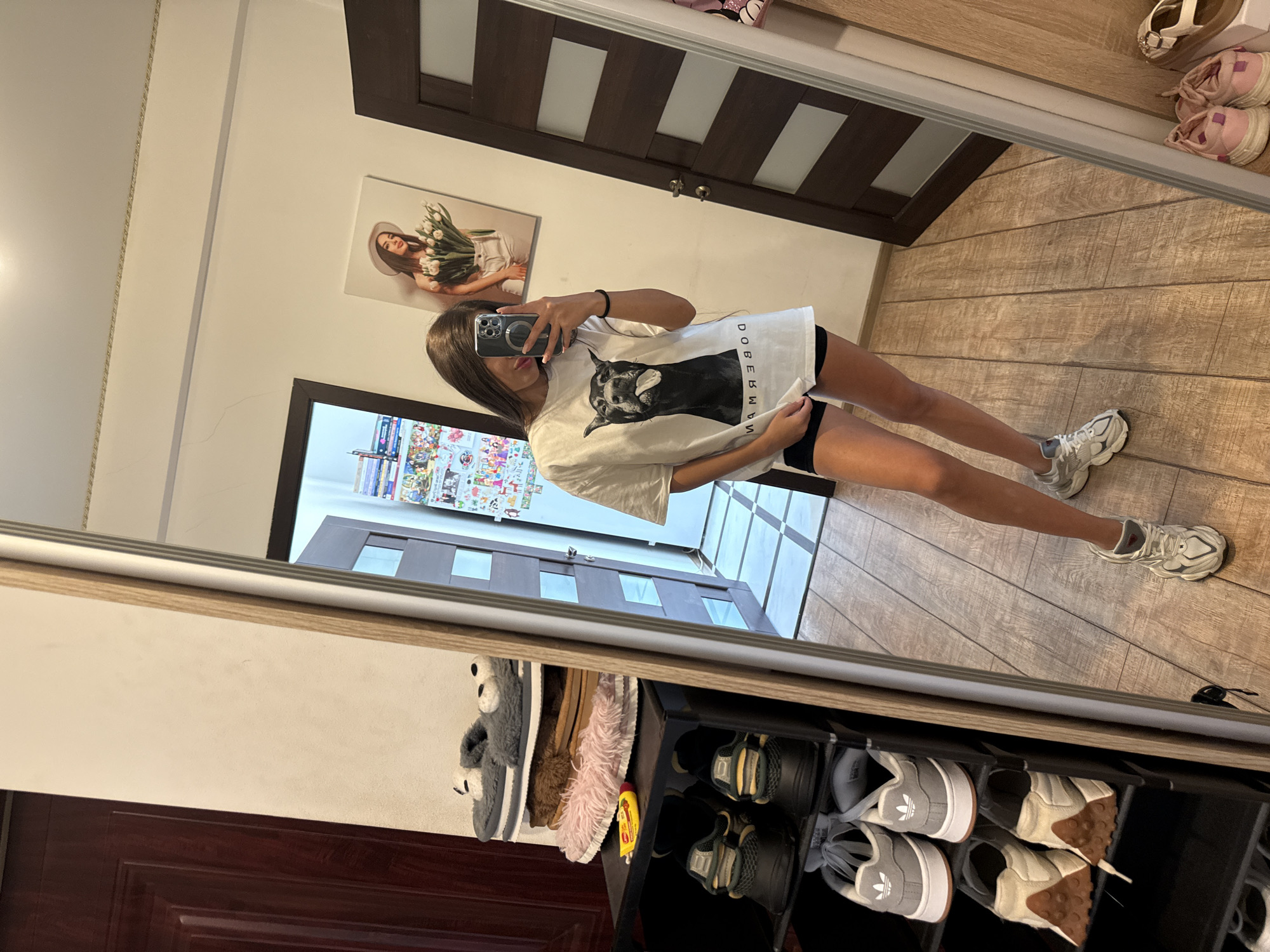 A young woman with long brown hair is taking a selfie in a full-length mirror in what appears to be a residential hallway or entryway. She is wearing a white t-shirt with a graphic print of a dog and black shorts. She is holding a smartphone in her right hand, angled to capture her reflection.
The mirror itself is framed in light wood and stands tall, reflecting a significant portion of the room. To the left of the mirror, a dark brown wooden door is visible. Beside the door is a black shoe rack filled with various pairs of shoes, including sneakers and slippers. On a shelf above the shoe rack, there are more slippers and a small yellow tube.
In the reflection of the mirror, a second dark brown wooden door is visible, partially open. On the wall to the left of this door, there is a framed picture of a woman holding a bouquet of tulips, wearing a wide-brimmed hat. Further to the left in the reflection, a collage of colorful images and text is attached to the wall. The floor is covered with light brown wooden planks. On the right side of the mirrored reflection, a portion of a tall wooden cabinet or closet is visible. Below this, on the floor, there are more shoes, including pink sneakers and beige sandals.
The setting is a domestic interior. Based on the provided information about the city, the location is assumed to be Chișinău, Moldova.