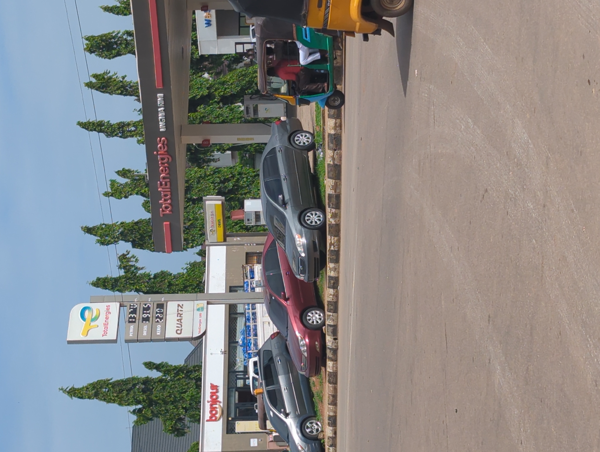 A medium shot shows the TotalEnergies petrol station at Unguwar Rimi, Nigeria, on a sunny day. The station has a red and white roof supported by white pillars. A prominent TotalEnergies sign with a yellow and blue logo stands tall, displaying fuel prices: PETROL 13.70, DIESEL 19.5, KERO 23.0, and QUARTZ. To the left of the sign, a building with "Bonjour" written in red letters is visible, with bottles of water stacked in the background. Several cars are parked at the station: a grey hatchback, a maroon sedan, and a dark grey sedan. A blue and green auto-rickshaw with two passengers, one wearing a red shirt and the other a white shirt, is parked in front of the station. A yellow auto-rickshaw is partially visible on the right side of the frame. The ground is paved with asphalt, and a line of decorative concrete blocks separates the road from the station. Tall, thin trees line the background, their green foliage contrasting with the clear blue sky.