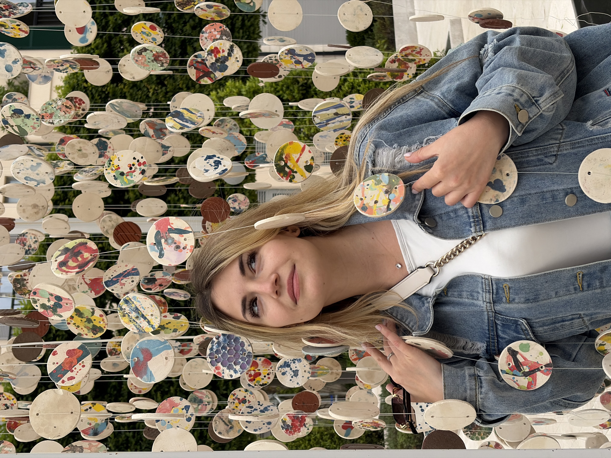 A young woman is posed among a display of painted ceramic disks in Tivat, Montenegro. She wears a denim jacket and a white top, and has light brown hair. The disks hang on strings, creating a visual barrier. The background shows green foliage and building exteriors, indicating an outdoor, urban setting.