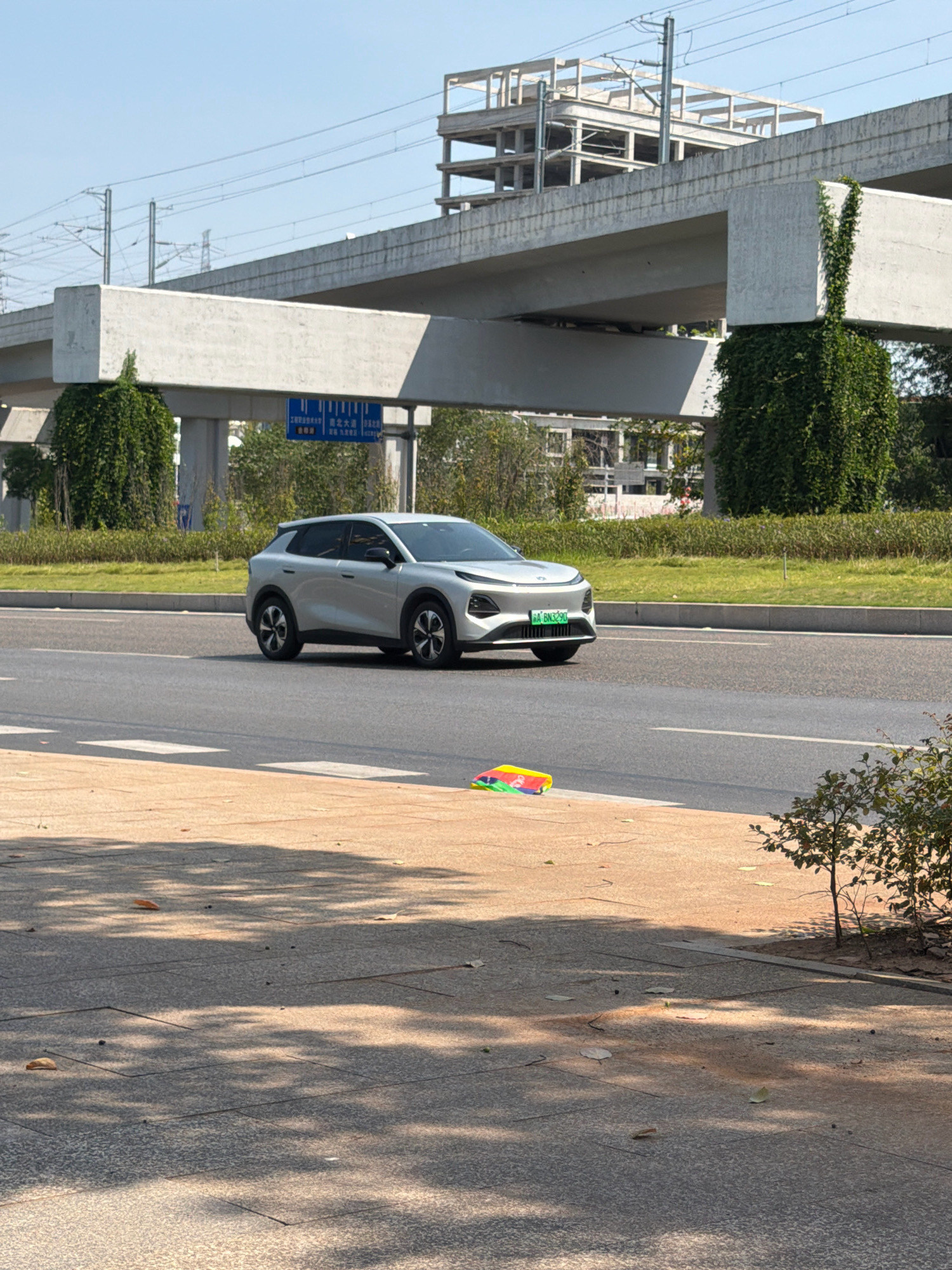A silver electric car, a BYD Yuan UP with license plate "A 6N3290", is traveling on a multi-lane road under an elevated highway structure. The highway pillars are covered in lush green ivy. Construction is visible in the background on the elevated structure. A blue street sign with Chinese characters is present on the side of the road. In the foreground, on the sidewalk, there is a colorful, flat object, possibly a toy or a piece of folded fabric. The ground is paved with concrete and shadows from trees are cast upon it. The sky is clear and blue.