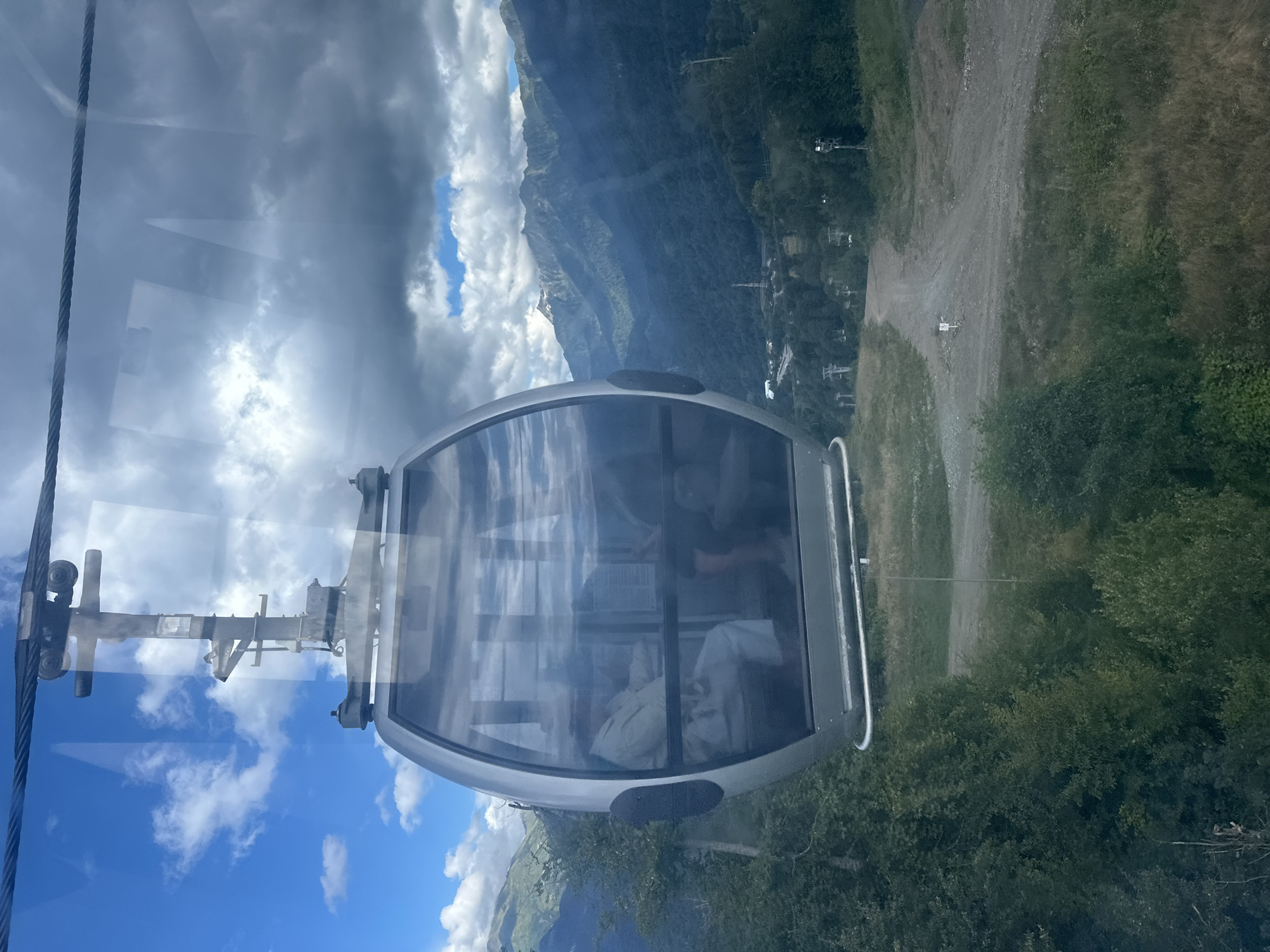 The image shows a cable car cabin suspended by a cable, moving forward through a mountainous landscape. The cabin is oval-shaped with large glass windows, revealing passengers inside. The environment is dominated by lush green trees and hills, with a dirt road visible below. In the distance, a range of mountains stretches across the horizon, partly shrouded in clouds. The sky is a mix of blue with scattered white clouds. The scene is set in Estosadok, Russia, a location known for its mountainous terrain and ski resorts. The cable car appears to be in motion, as indicated by the slight blur in the reflection on the cabin windows and the perspective of the surrounding environment. The overall impression is one of travel and scenic views in a natural, mountainous setting.