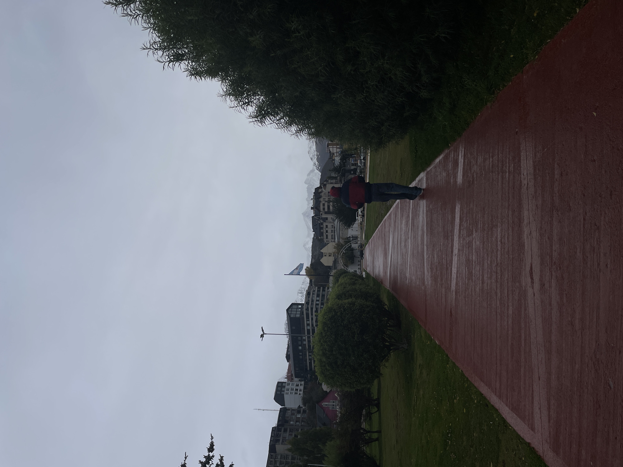 An outdoor shot captures a person walking along a red track in Ushuaia, Argentina. The sky is overcast. In the background, the city's buildings rise toward snow-capped mountains. A flag flies in the midground. Green grass and trees border the track. The photo is taken from a slightly elevated perspective.