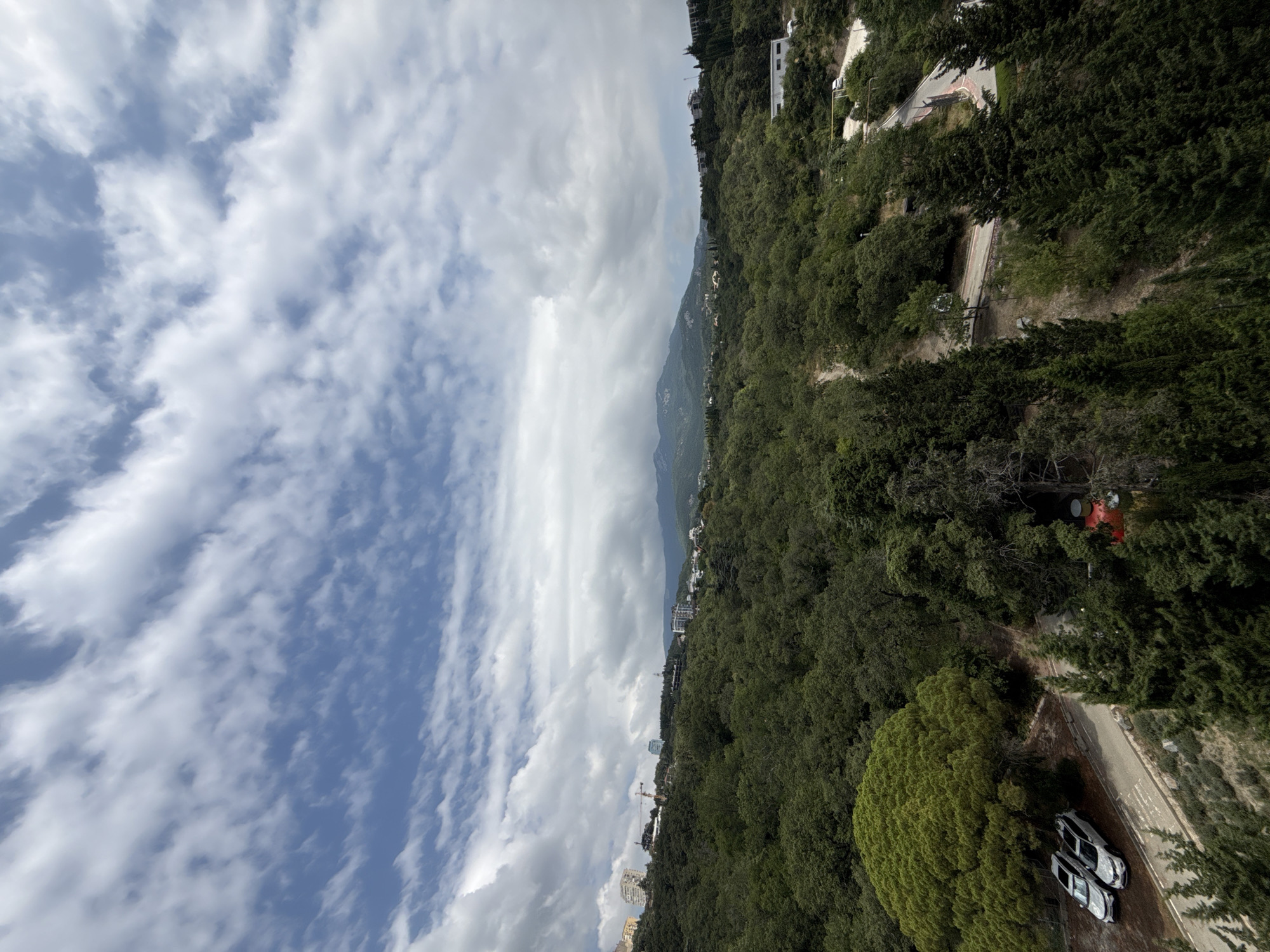 The image shows a panoramic view of a forested hillside with a town or city built on its upper slopes, overlooking a valley. The sky is filled with dynamic white and gray clouds against a bright blue sky. The hillside is densely covered with various types of trees, predominantly dark green conifers and lighter green deciduous trees.
In the foreground and midground, winding roads are visible, with a few cars parked on one of them. To the left, two silver cars are parked on a paved area next to a large, bright green, bushy tree. Further up the hillside, buildings of various sizes and architectural styles are scattered amongst the trees, suggesting a residential or resort area. Some of these buildings appear to be modern apartment complexes, while others are smaller structures. Construction cranes are also visible in the distance, indicating ongoing development.
The background features rolling hills and distant mountains, their shapes softened by atmospheric haze. The overall impression is of a verdant, mountainous landscape with human settlement integrated into its natural contours. The scene appears to be captured from an elevated viewpoint, possibly a balcony or a higher point on the hillside.