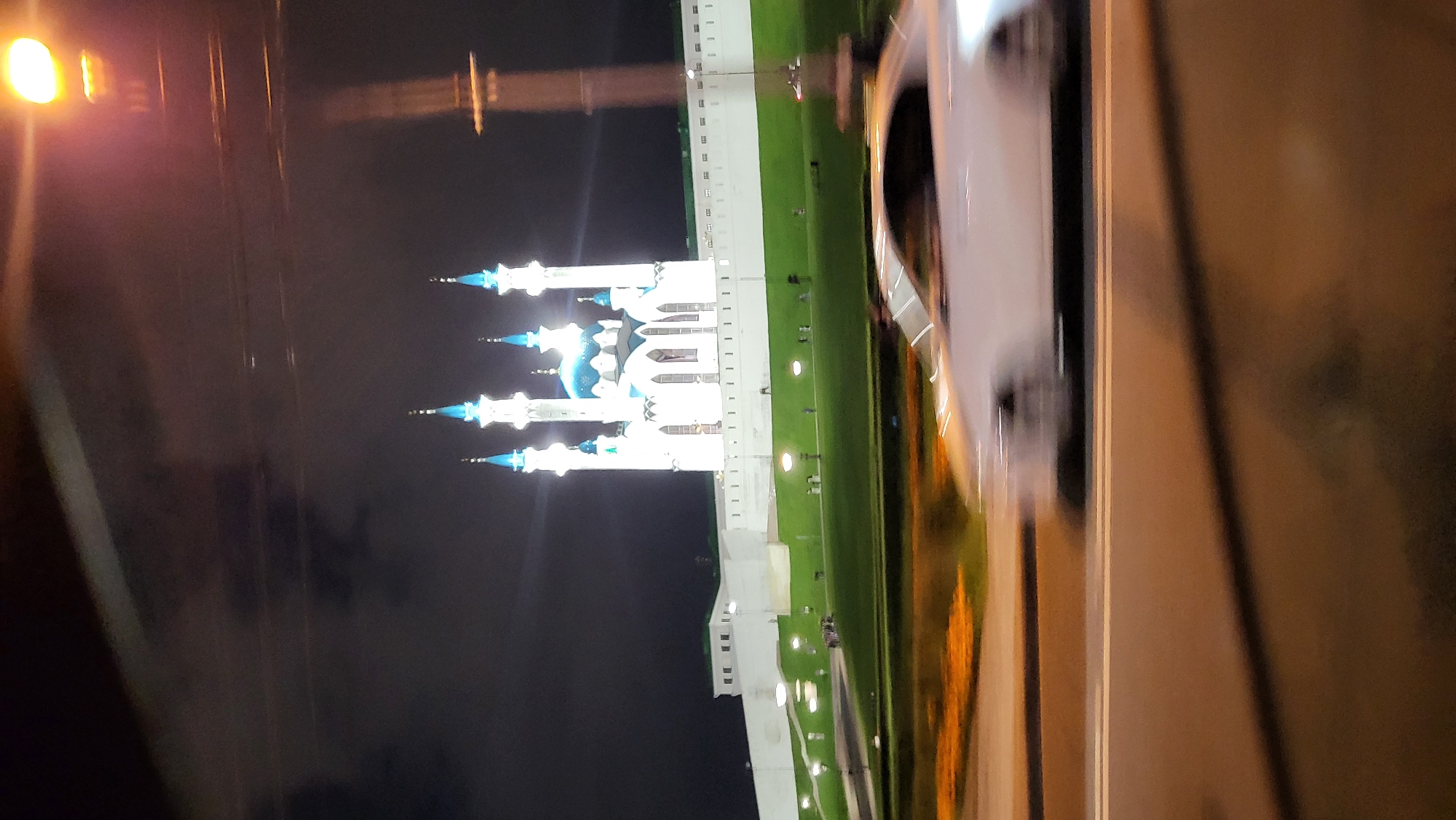 The image shows a night view of the Kul Sharif Mosque in Kazan, Russia. The mosque, with its white walls and blue domes and minarets, is illuminated against the dark sky. In the foreground, a white car is seen in motion, blurred due to the camera's movement. The car is on a road, and there is a grassy area with lights and some people visible between the road and the mosque. The overall scene suggests a passing shot taken from a vehicle.