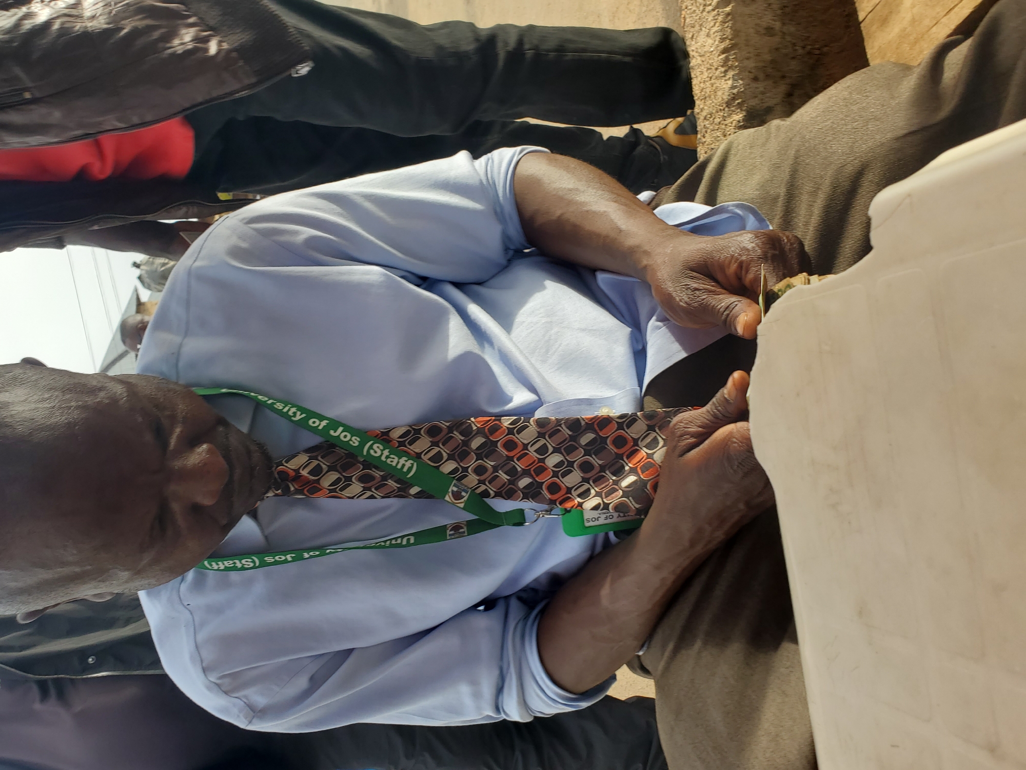 This media file shows a close-up, rotated view of two individuals, with one person in the foreground wearing a light blue collared shirt and a patterned tie. This individual has dark skin and appears to be focused on something held in their hands. A green lanyard with visible text is around their neck, with the words "University of Jos (Staff)" discernible. The background shows parts of other people's clothing, including dark pants and a red garment, suggesting a gathering or public space. The lighting indicates it is daytime. The scene appears to be outdoors, possibly in an urban or semi-urban environment, given the rough texture of a wall or ground in the background. The overall impression is that of an ordinary day, perhaps at an event or during a break, with the focus on the person in the foreground and their activity.