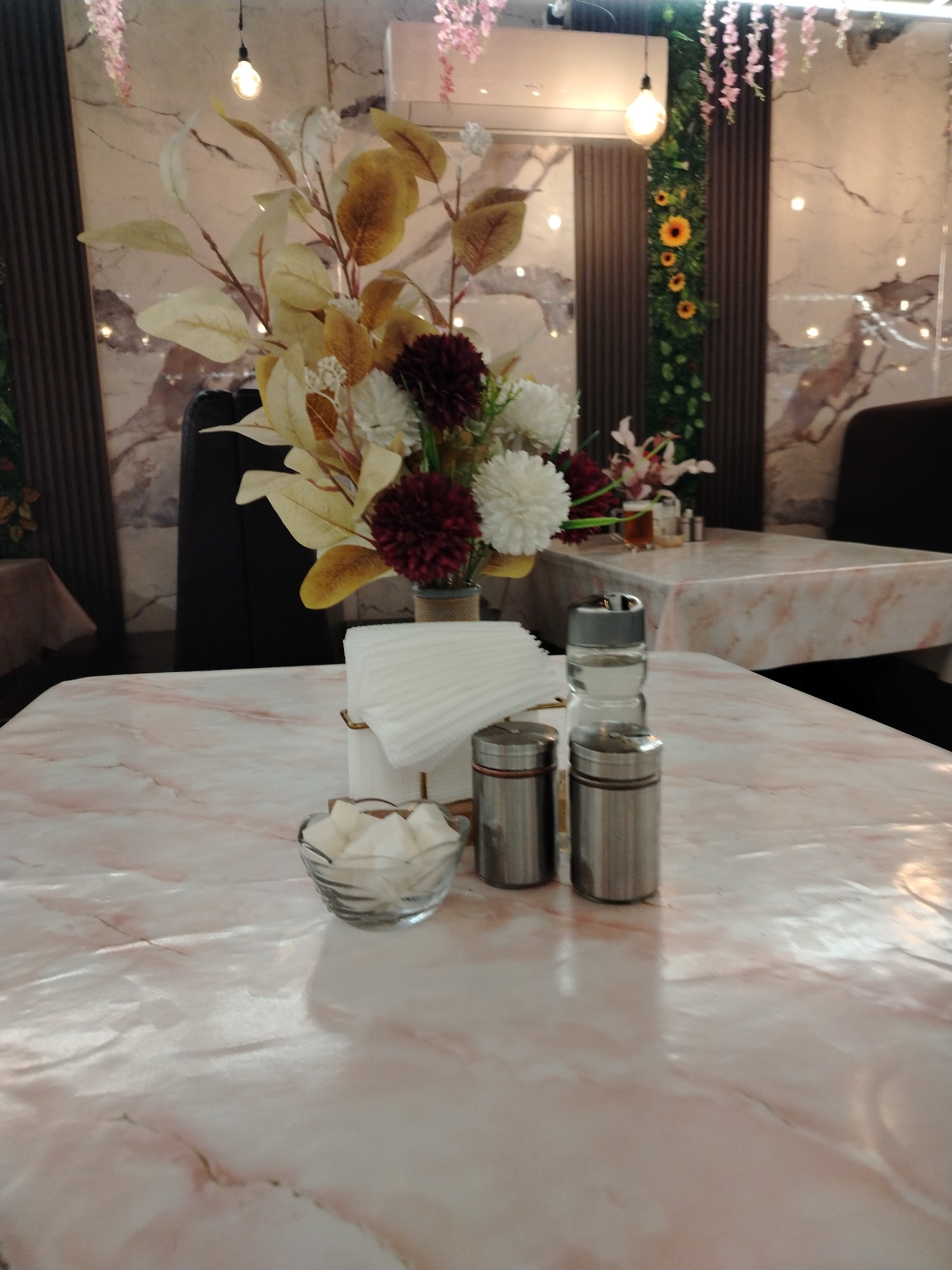 The image shows a close-up view of a table setting in what appears to be a restaurant or cafe. The table has a pink and white marble-patterned tablecloth. On the table are several items: a small glass bowl filled with white cubes, possibly sugar or artificial sweetener; two silver cylindrical containers, likely salt and pepper shakers; and a clear glass bottle with a silver cap, possibly a water dispenser or a decorative element. Behind these items, a bouquet of artificial flowers in various shades of white, burgundy, and yellow is arranged in a small vase.
In the background, a large artificial floral arrangement is visible, with some pink flowers hanging from above. The wall behind the arrangement has a marble-like pattern. There are also vertical dark brown panels and some greenery with yellow flowers. A light fixture with a single bulb hangs from the ceiling. Another table with a similar tablecloth is visible in the background, with a glass of amber liquid and some small containers on it. The overall impression is one of a decorated indoor space, possibly a dining area. There are no people visible in the image. The lighting suggests it could be daytime or an indoor setting with artificial lighting.