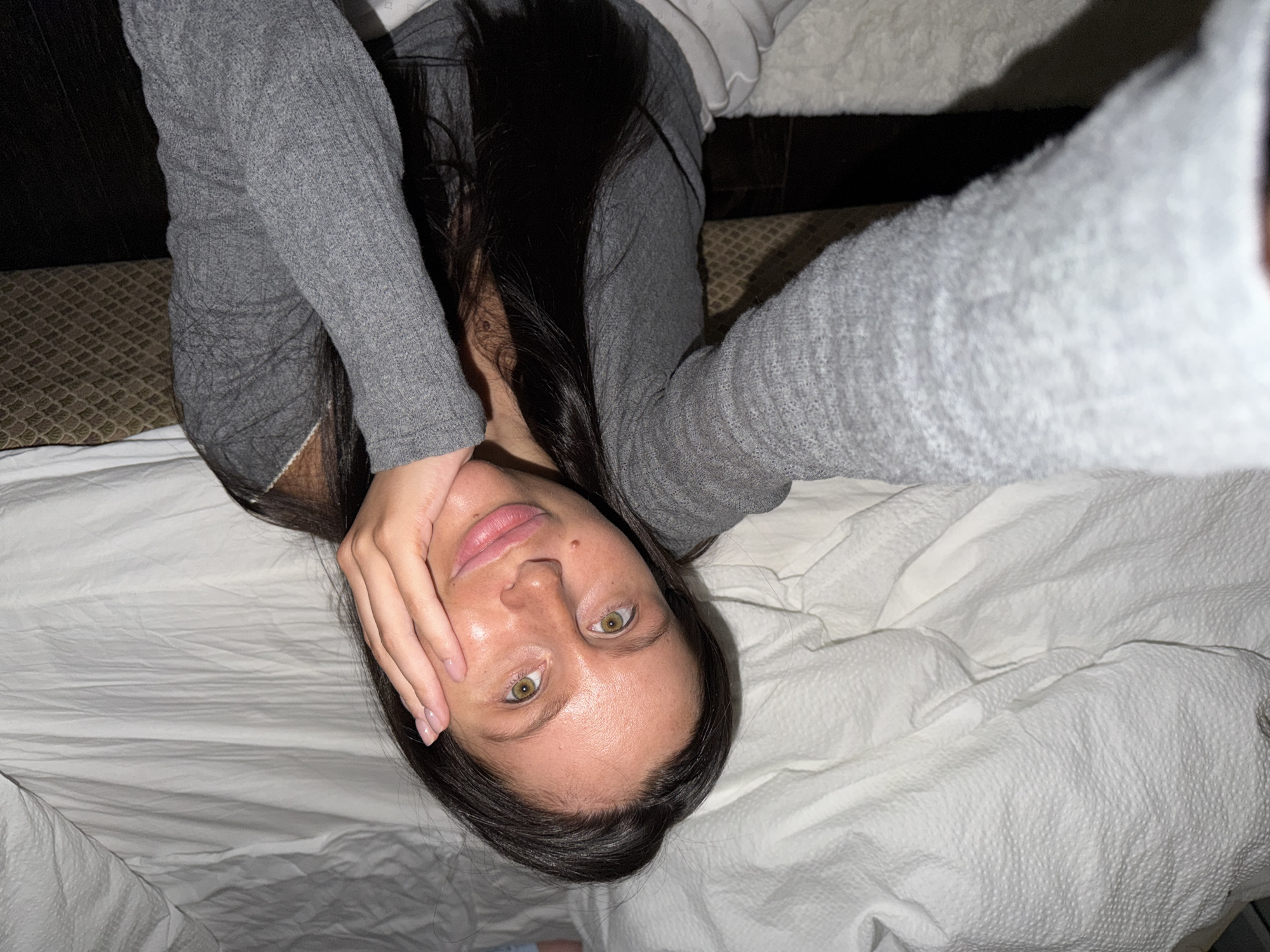 A young woman with dark hair is lying on a white bed. She is wearing a gray long-sleeved shirt. Her face is visible, with light eyes and fair skin. One hand is resting on her cheek. The background is blurred, but a dark headboard is visible. The location is not visible from the image, and it is impossible to determine if the image was taken in Khimki, Russia.