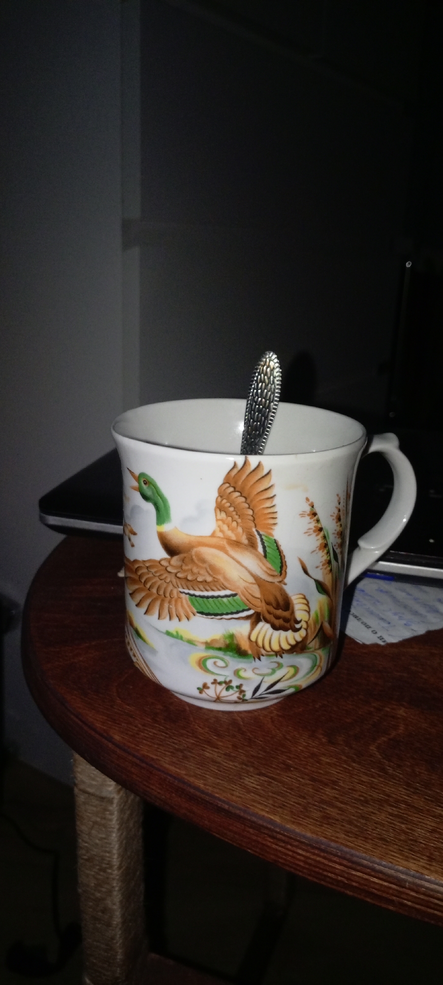A close-up, low-angle shot shows a white ceramic mug adorned with a painted scene of two mallard ducks in flight against a pale background. One duck is in the foreground, with its green head and brown and orange wings spread, while another is partially visible behind it. A silver spoon with a textured handle is placed inside the mug. The mug sits on a dark brown, wooden table with a visible grain pattern. In the background, a dark surface, possibly a laptop, is partially visible, and behind that, a dark, out-of-focus environment suggesting an indoor setting. The lighting is dim, with shadows obscuring details in the background. The scene appears to be indoors, possibly in a home environment. There is no indication of the specific location within the city of Зюзино, Russia.
