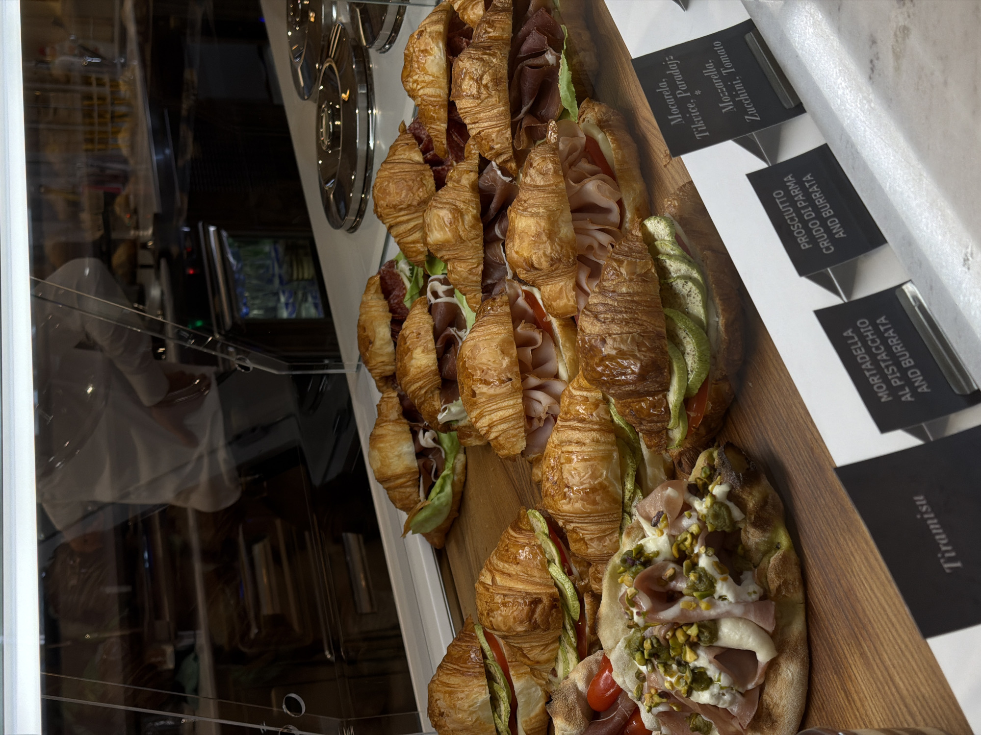 The image captures a display of gourmet sandwiches, likely inside a deli or café, possibly in Budva, Montenegro, given the context provided. The sandwiches are predominantly croissant-based, filled with various combinations of ingredients, including prosciutto, mozzarella, zucchini, and tomato. One particular sandwich or flatbread variation features mortadella, pistachio, and burrata. Small black informational cards next to the items detail the ingredients in each sandwich. The sandwiches are displayed on a wooden surface behind a glass enclosure. Reflections are visible in the glass.