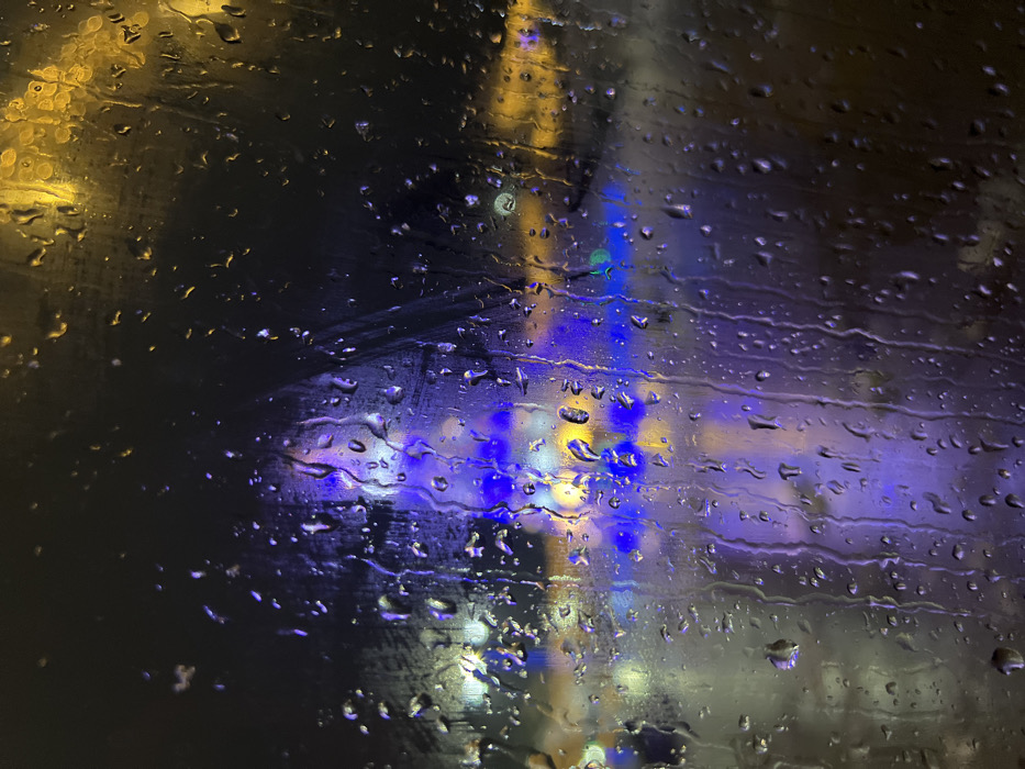 The image depicts a nocturnal scene in the city of Terjola, Georgia, observed through a transparent surface heavily covered in water droplets and vertical streaks of rain. The foreground is dominated by these liquid formations, varying in size and shape, indicating active precipitation. The background is significantly blurred, presenting an out-of-focus array of artificial lights. A tall, conical structure, predominantly illuminated by blue lights, occupies the central background, consistent with a festive light installation or Christmas tree. Multiple diffuse yellow and orange light sources are visible throughout the mid-ground and upper right, suggesting streetlights or building illuminations. Faint green and white light blurs are also present. The overall environment is dark, signifying nighttime, with the rain-streaked view obscuring specific details of the urban landscape. No human figures, vehicles, or explicit actions are discernible.
